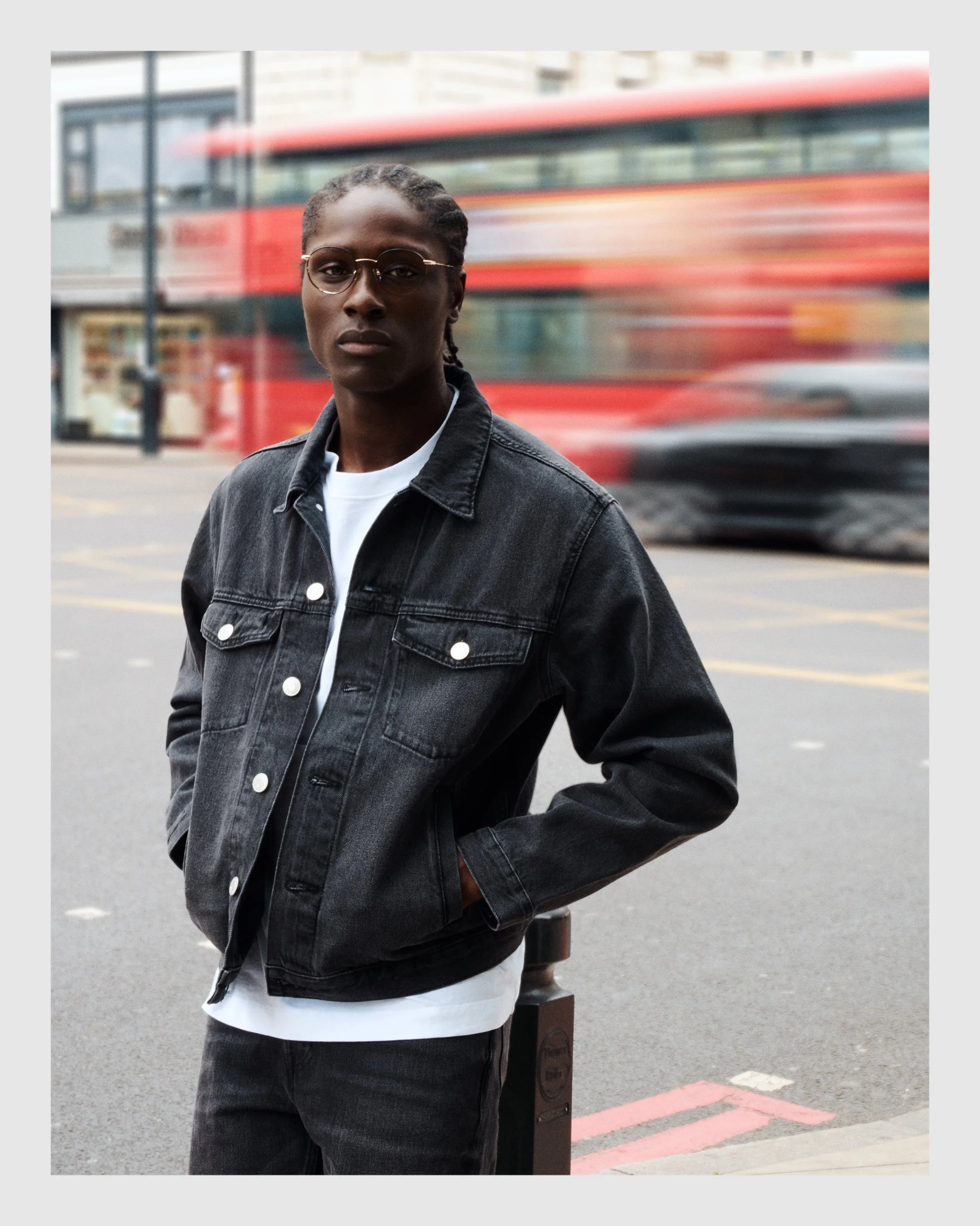 Male model in a black denim jacket wearing optical glasses with a red London bus passing behind him.