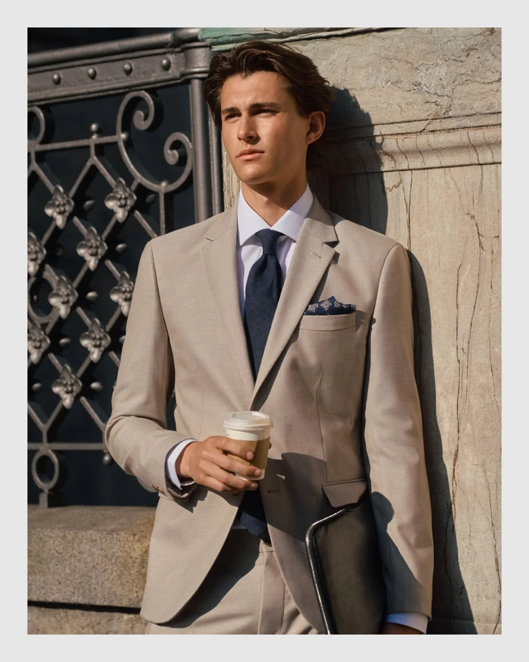 Male model wearing a beige Brothers suit with navy tie, holding a coffee and leather portfolio