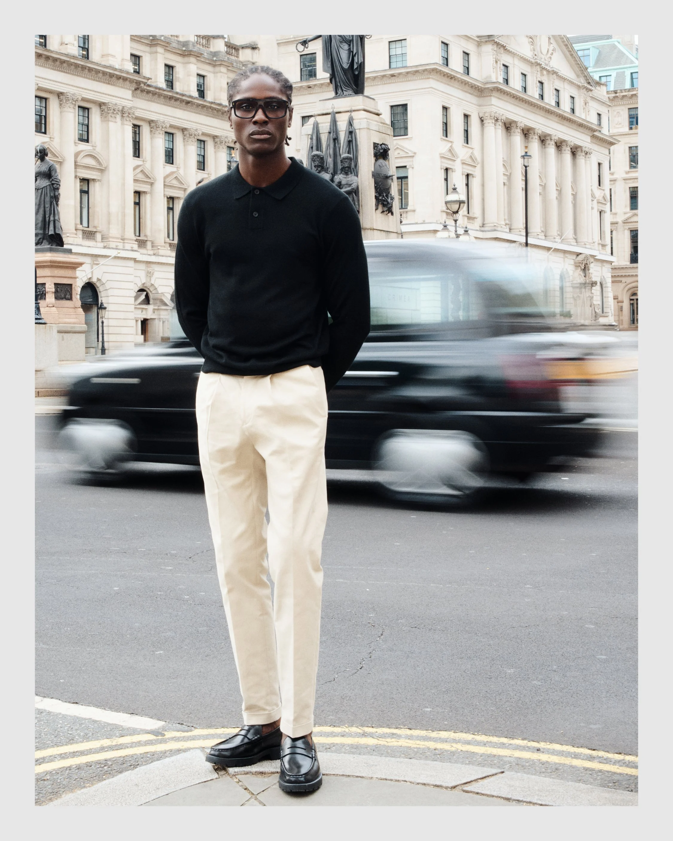 Male model wearing black sunglasses, a black polo shirt and light trousers with a moving London taxi behind him.