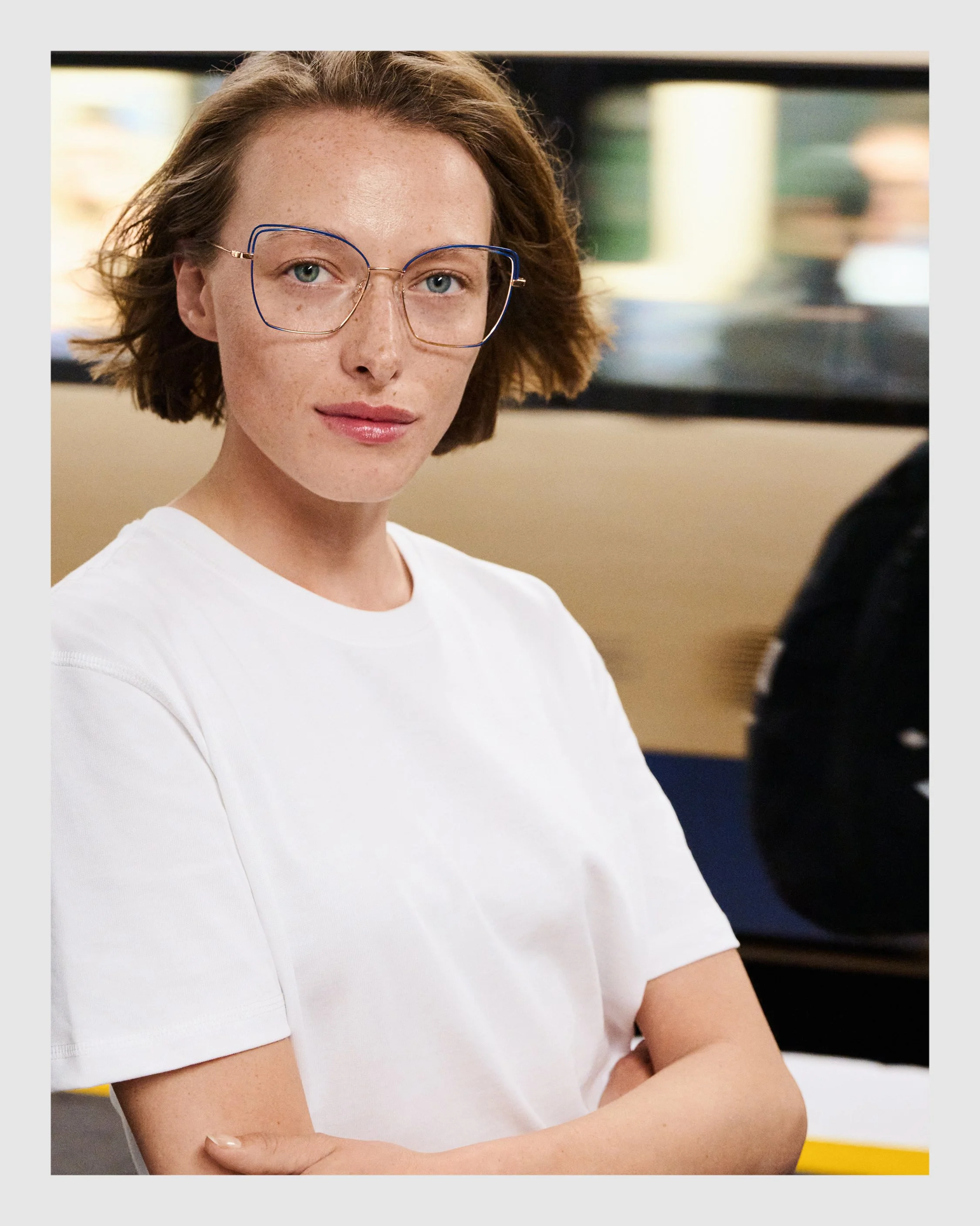 Female model wearing blue and gold optical glasses and a white T-shirt inside a London Underground train.