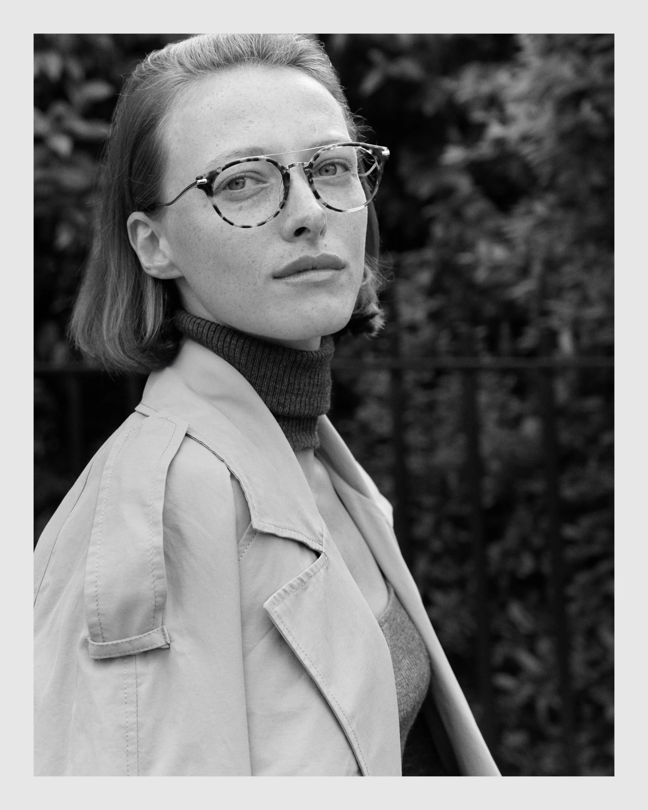 Black-and-white portrait of a female model wearing patterned optical glasses and a trench coat.