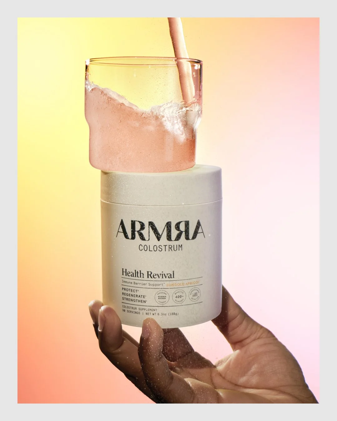 Hand holding ARMRA Health Revival product beneath a glass of pink drink