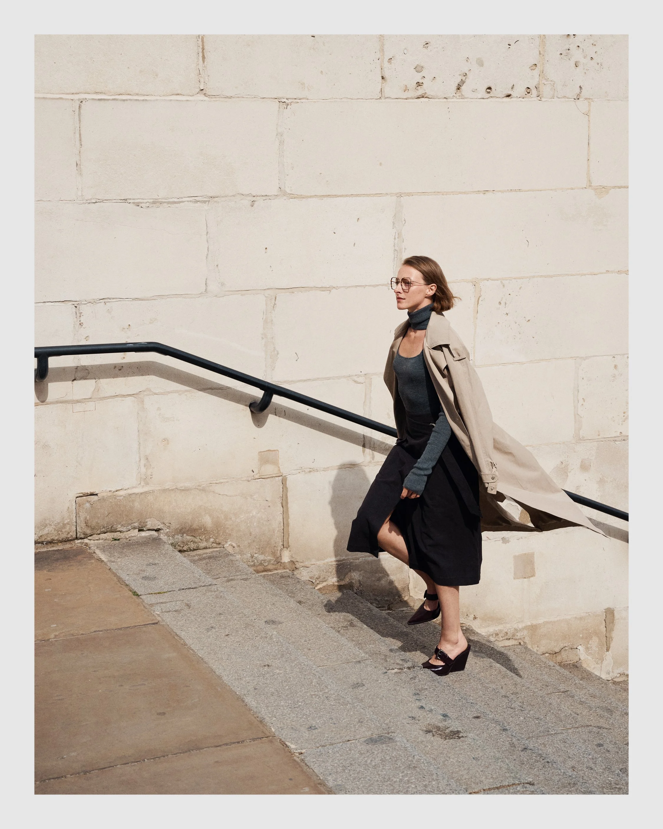 Female model in a trench coat and dark outfit walking beside pale stone architecture in London.