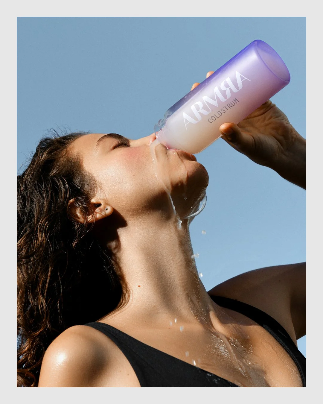 Woman drinking from a purple ARMRA colostrum bottle against a blue background