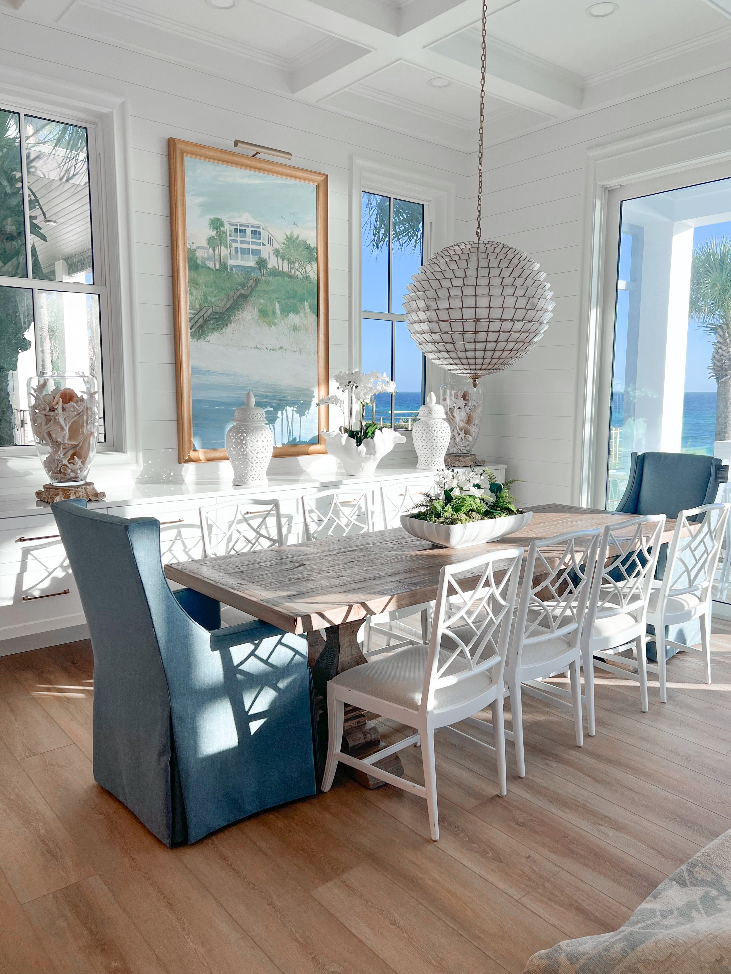 Dining room with a rustic wooden table surrounded by white chairs and a blue chair, decorative vases, a painting of a beach house, large windows with an ocean view, and a sphere-shaped chandelier.