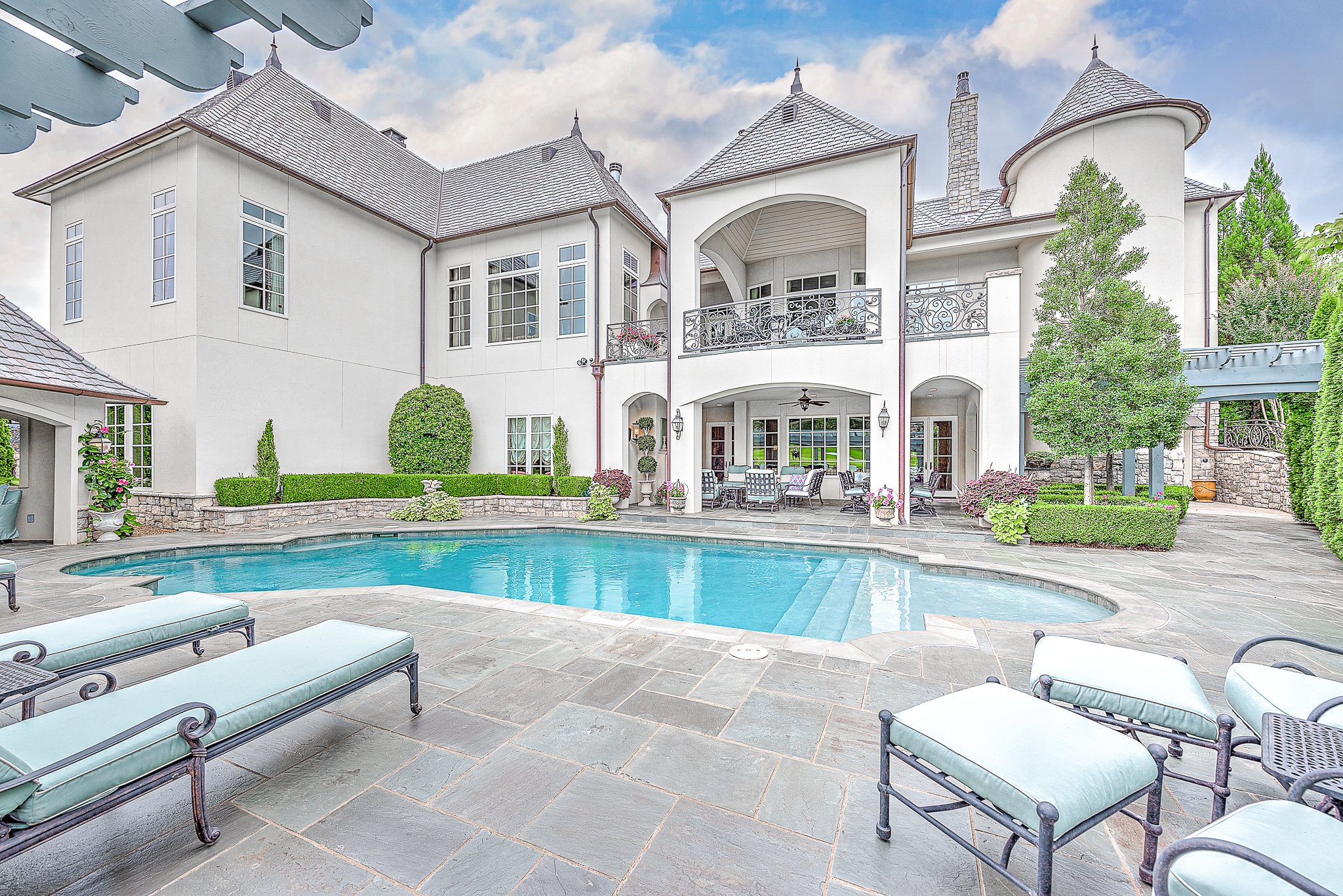 Luxurious mansion with a swimming pool, patio furniture, and well-manicured landscaping under a clear blue sky.