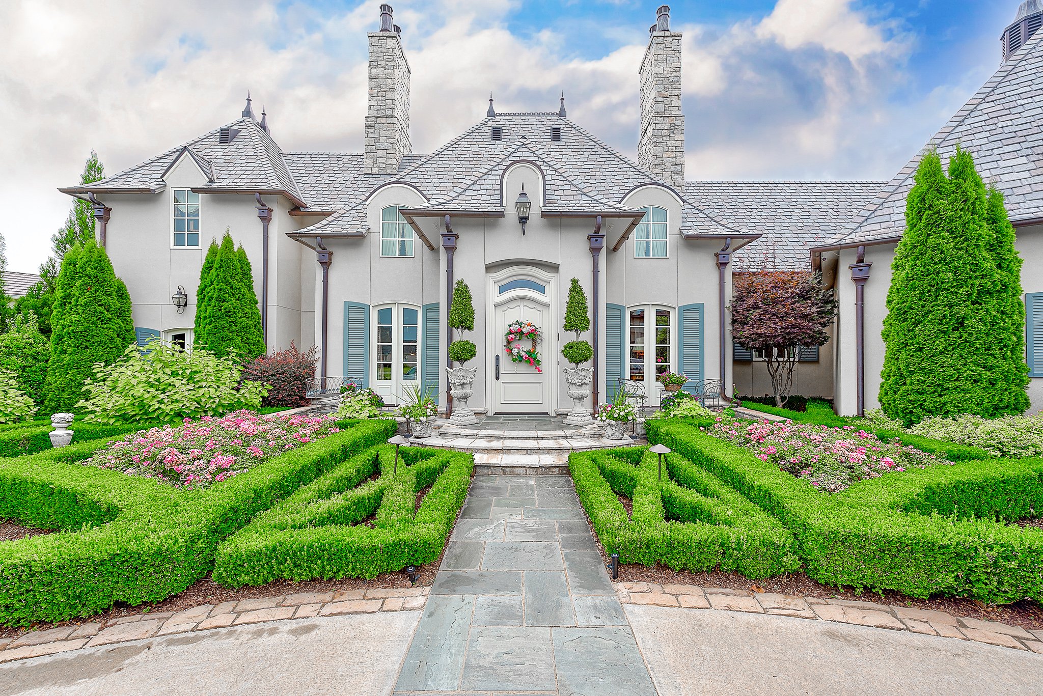 Elegant French-style mansion with ornate landscaping, featuring a symmetrical front garden with trimmed hedges and colorful flowers. The house has a stone facade, multiple chimneys, blue shutters, and a grand entrance with columns and decorative urns