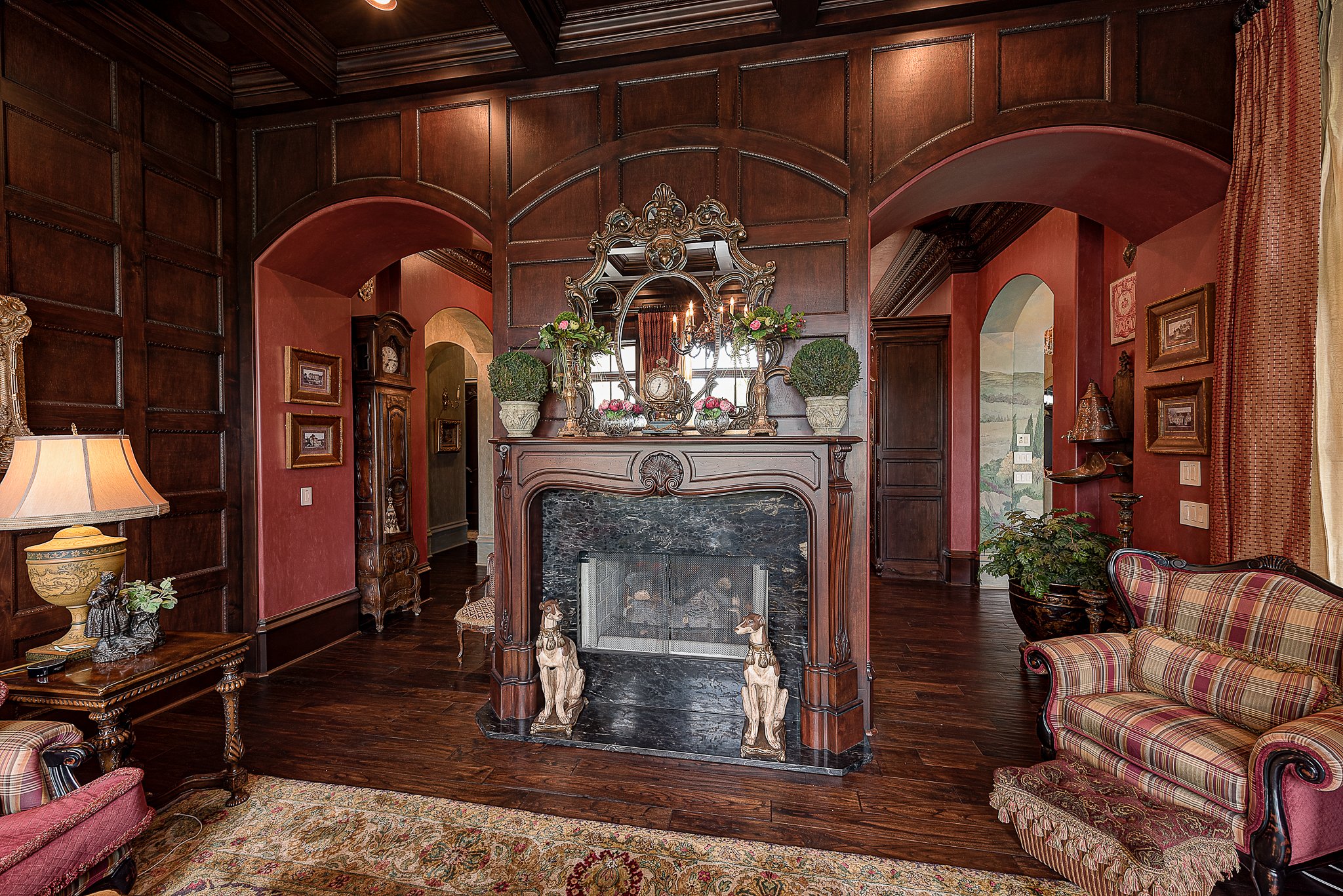 Elegant living room with dark wood paneling, ornate fireplace, antique furnishings, floral decorations, and patterned curtains.