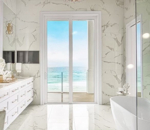 Luxury bathroom with marble walls, double vanity, freestanding tub, and sea view through glass doors.
