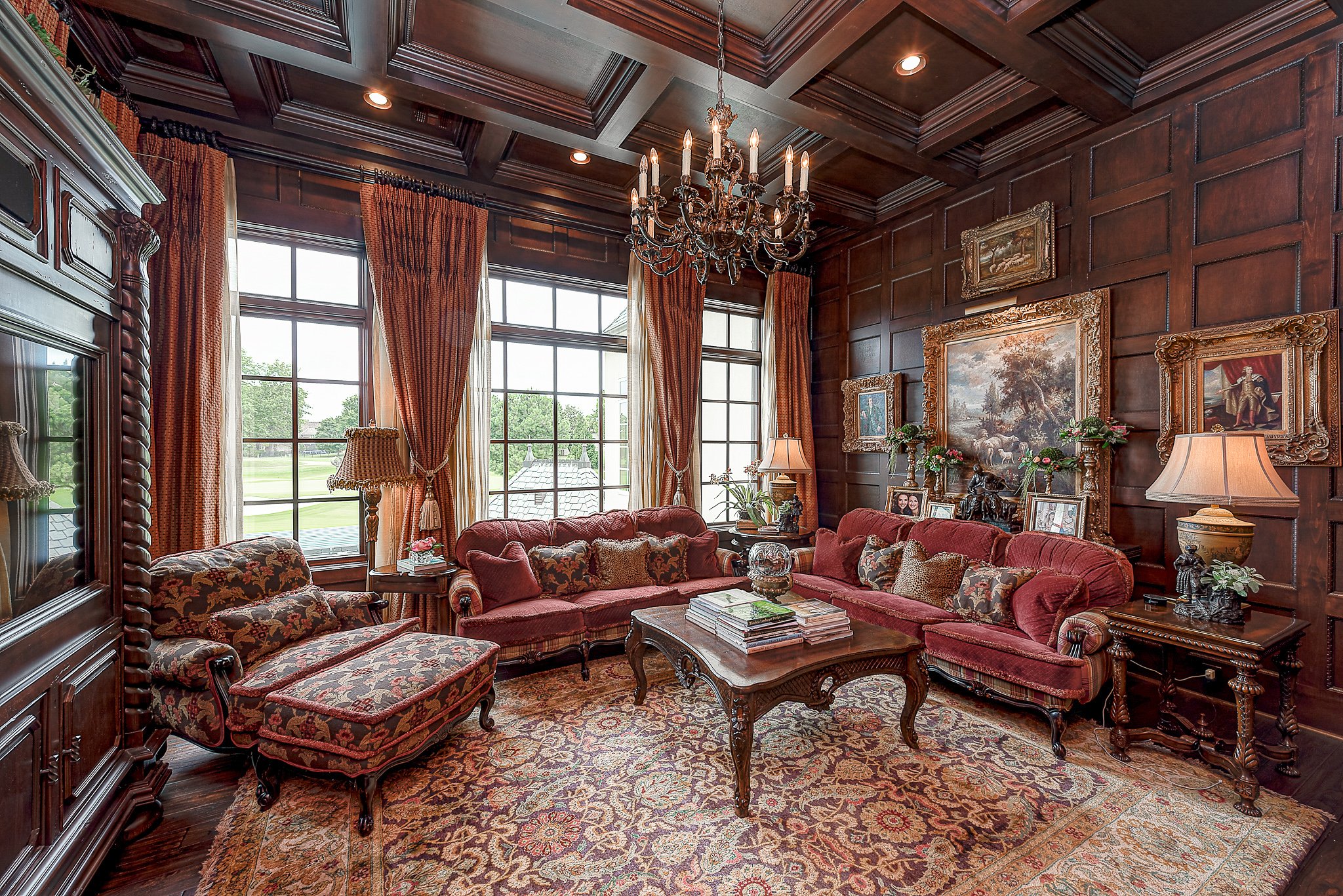 Luxurious living room with vintage furniture, ornate chandelier, wood paneling, floral-patterned upholstery, and large framed paintings. Large windows with heavy drapes allow natural light. A rich rug covers the floor, and there are decorative lamps