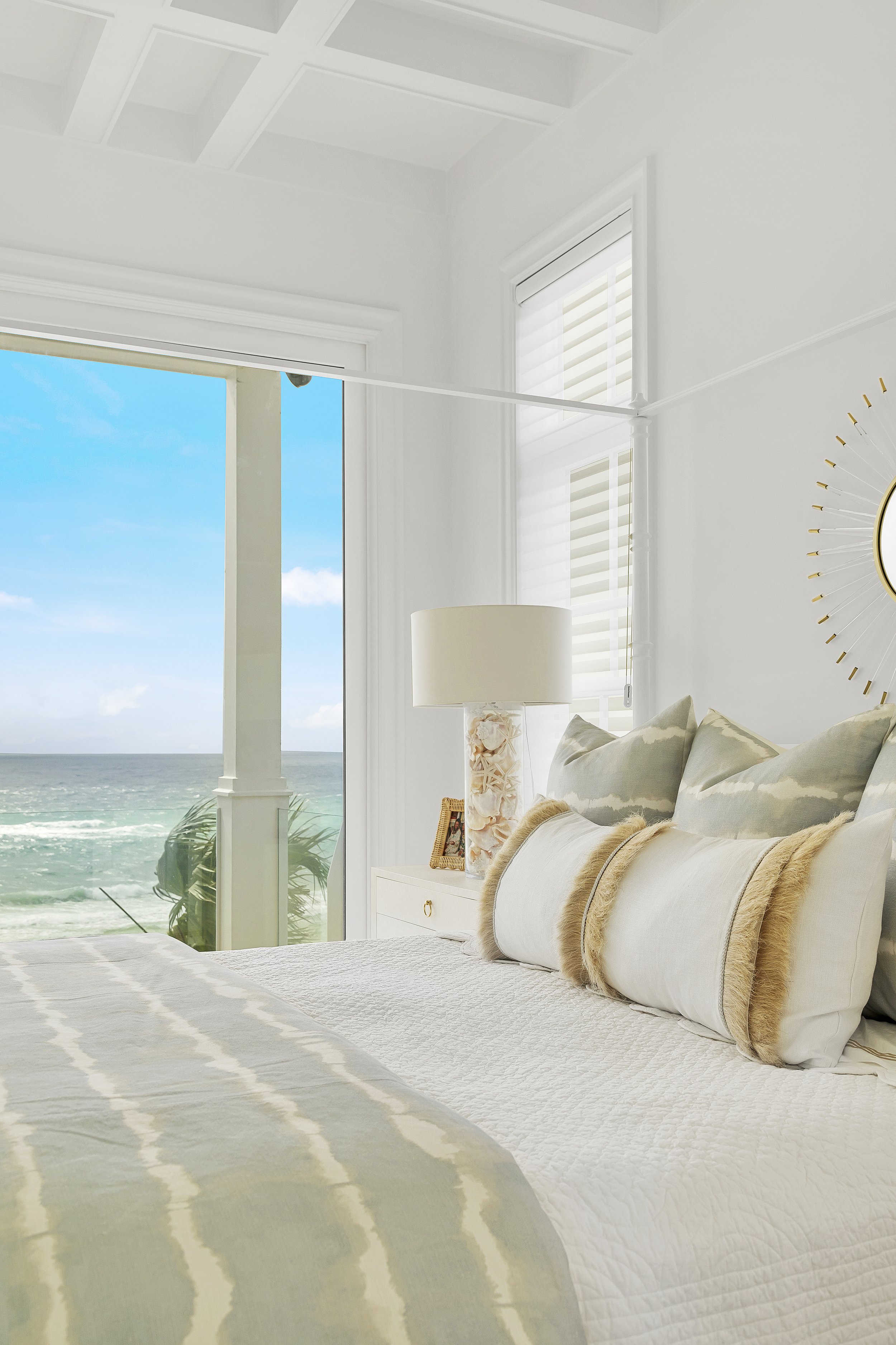 Beachside bedroom with ocean view, sunburst mirror, shell lamp, and decorative pillows.