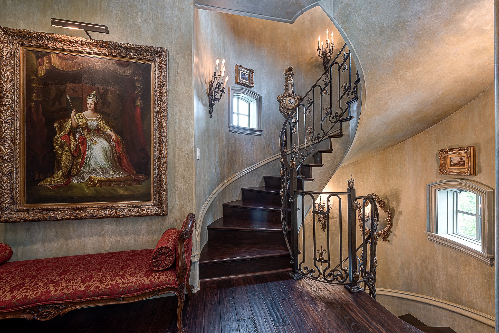 Elegant interior spiral staircase with ornate iron railing, vintage painting of a royal figure on the wall, decorated with opulent lighting, and a red upholstered bench.