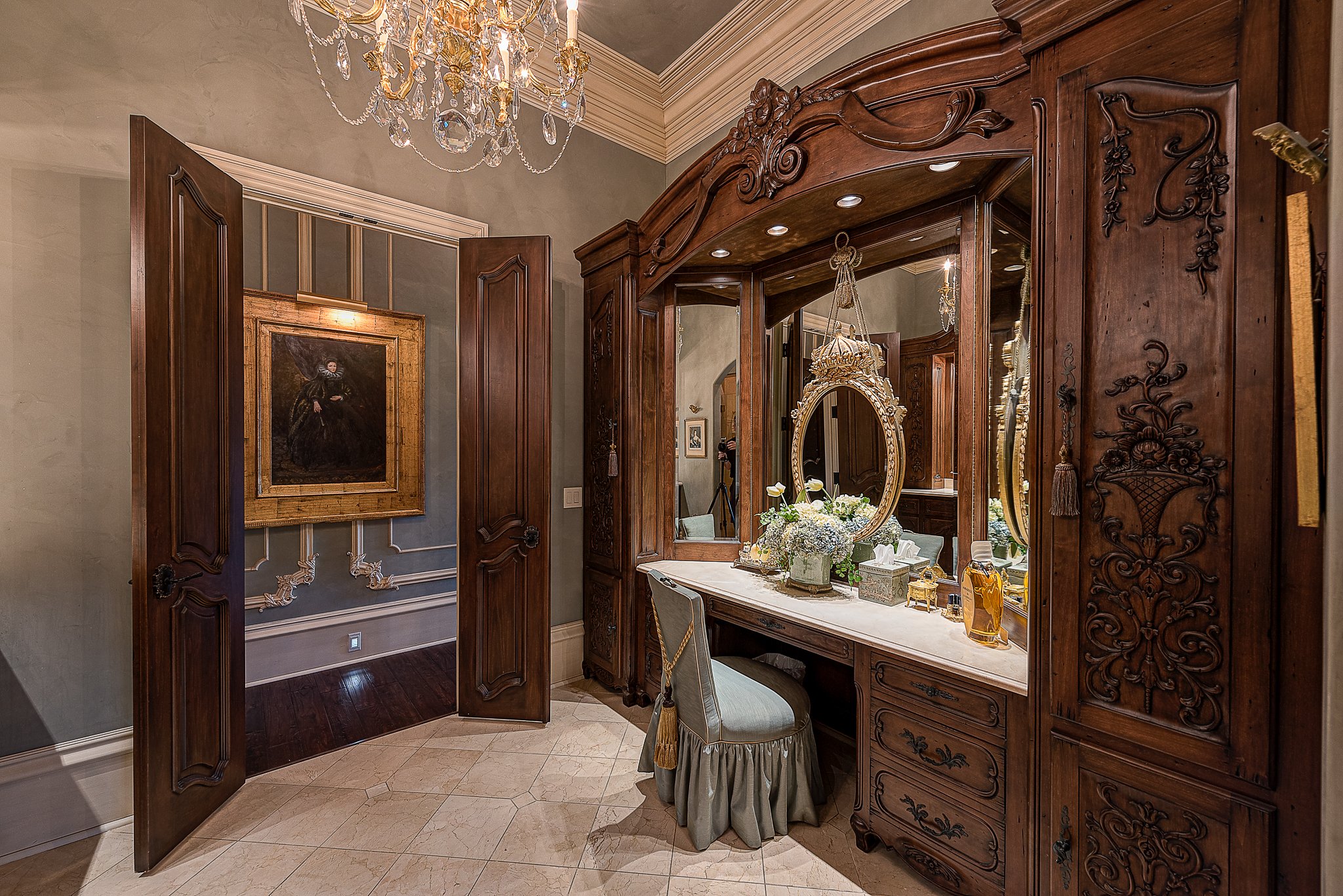 Elegant dressing room with ornate wood vanity, round mirror, and decorative chair, featuring a chandelier and a framed painting on the wall.