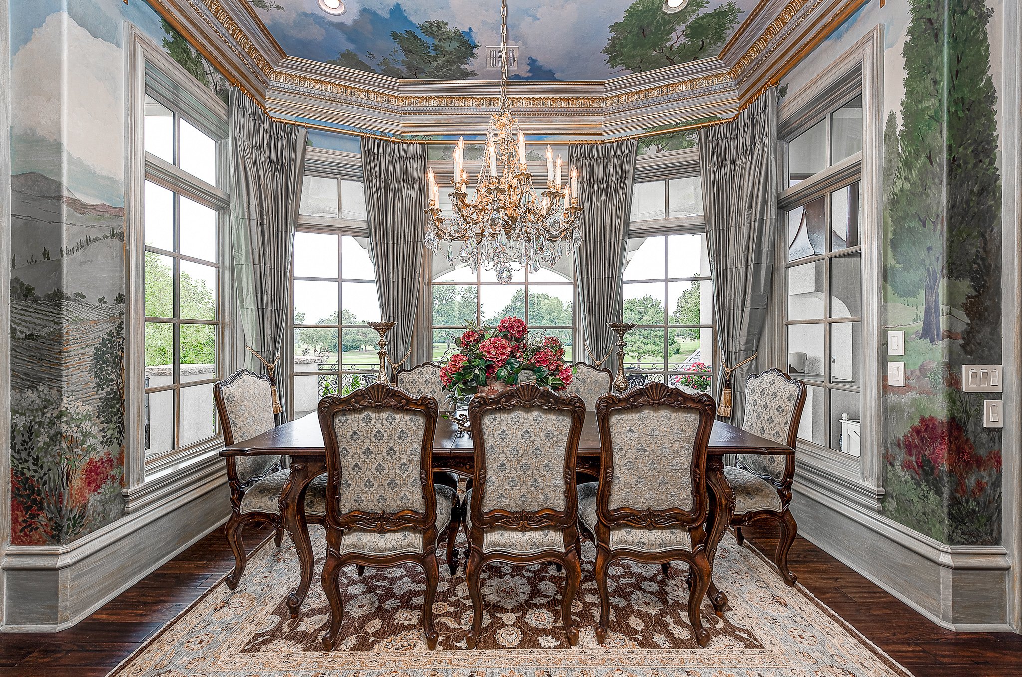 Elegant dining room with ornate wooden chairs arranged around a dark wooden table, a large chandelier overhead, landscape mural walls, and floor-to-ceiling windows with drapes.