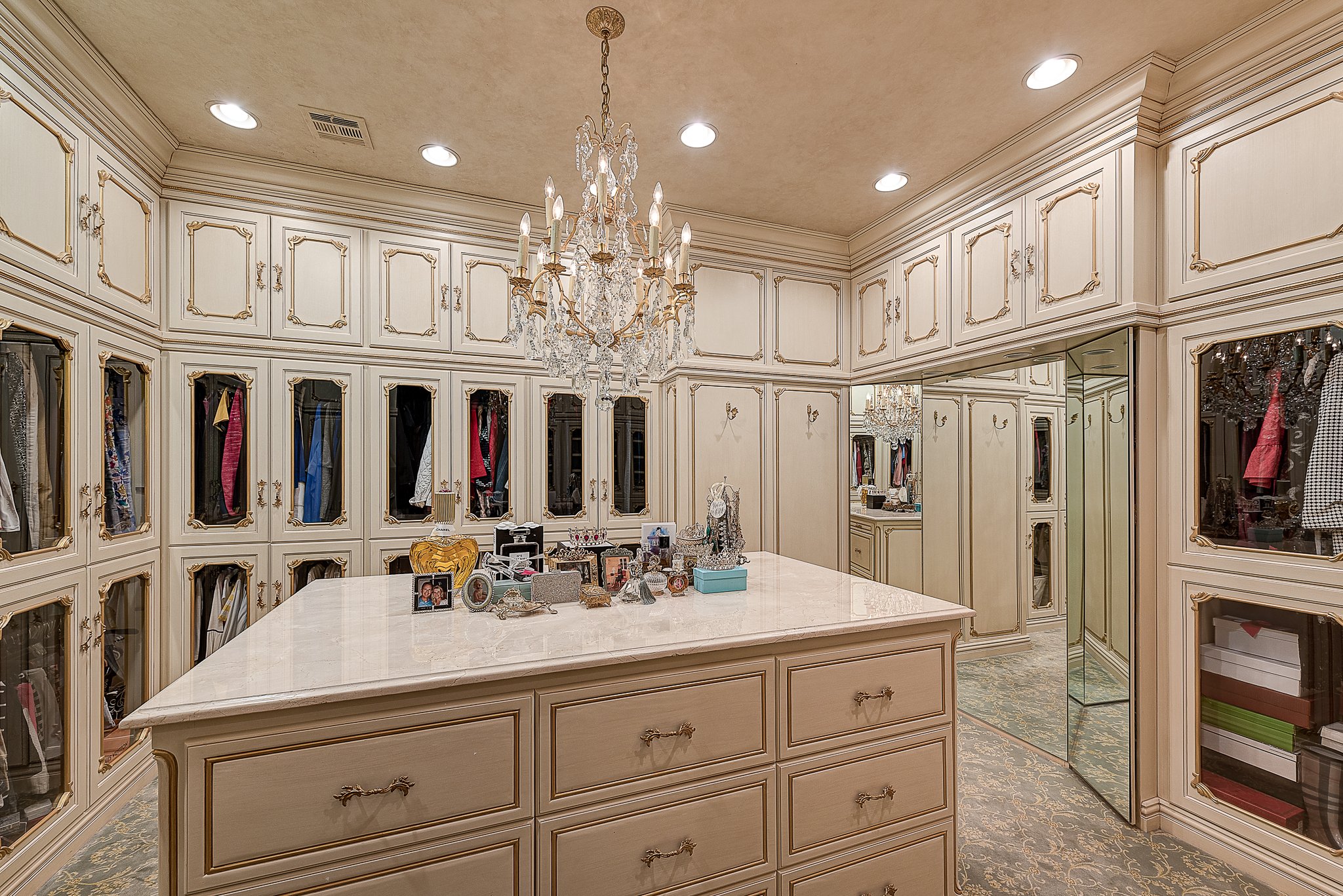 Luxury walk-in closet with chandelier, white cabinetry, mirrored doors, and clothing displays. Features a central island with jewelry and personal items on top.