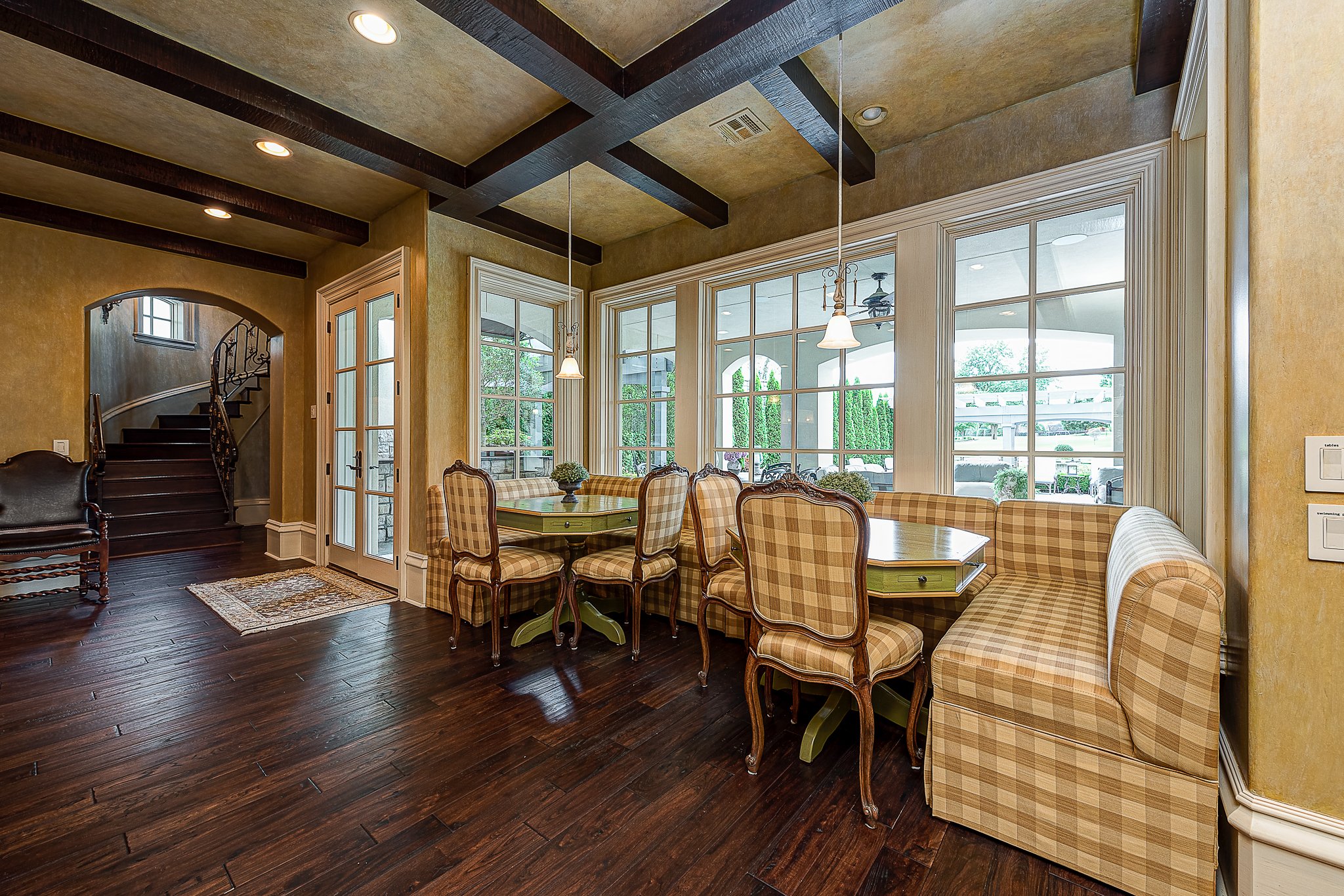 Dining area with plaid upholstery, wooden chairs, a green table, large windows, and dark wood floors next to a staircase in a house.