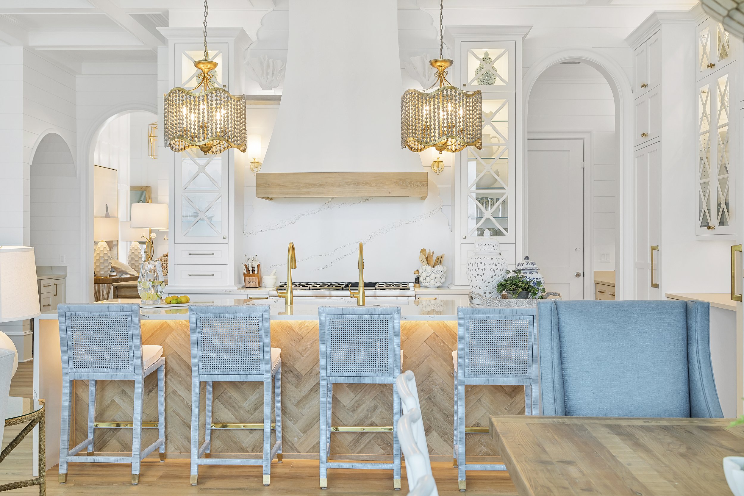 Modern kitchen interior with light blue barstools, gold pendant lights, wooden countertops, and white cabinets. Features a large range hood and decorative vases.