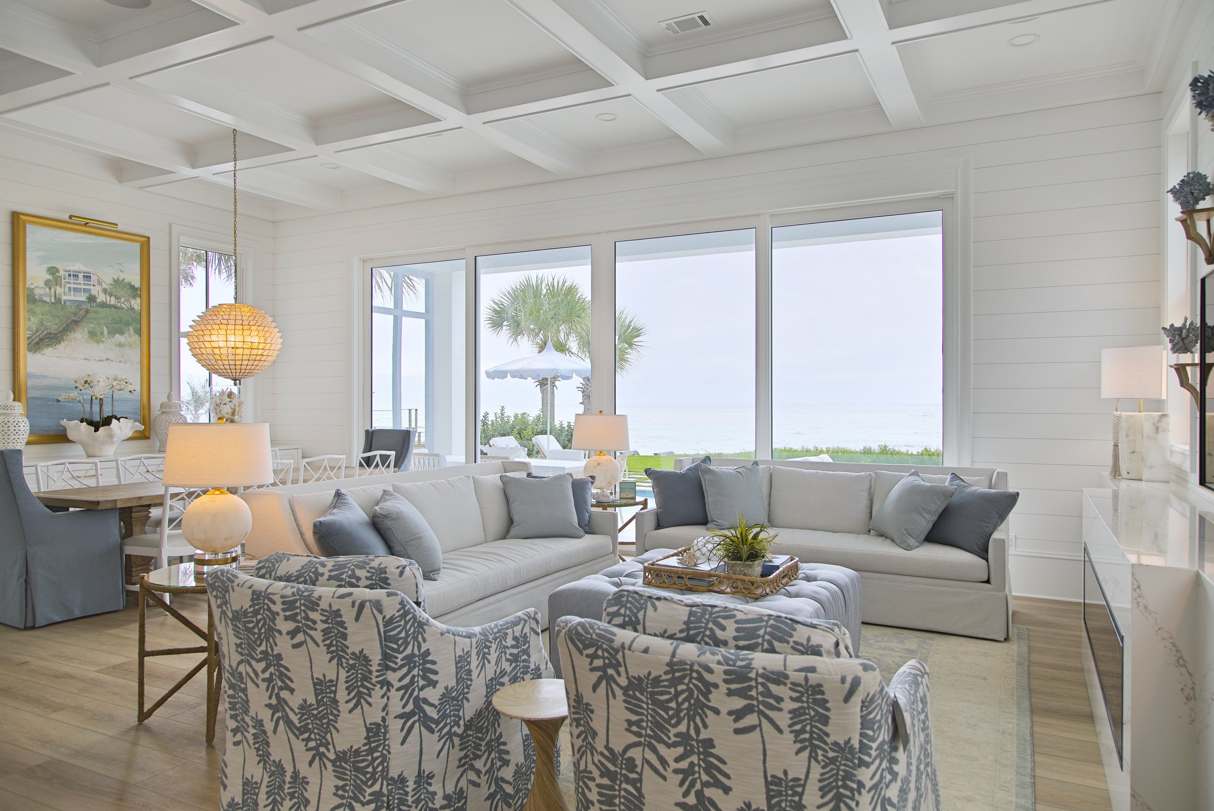 Coastal-themed living room with large windows overlooking the ocean, featuring light-colored sofas, patterned chairs, a wooden coffee table, and a pendant light. Tropical decor and artwork enhance the beachy atmosphere.