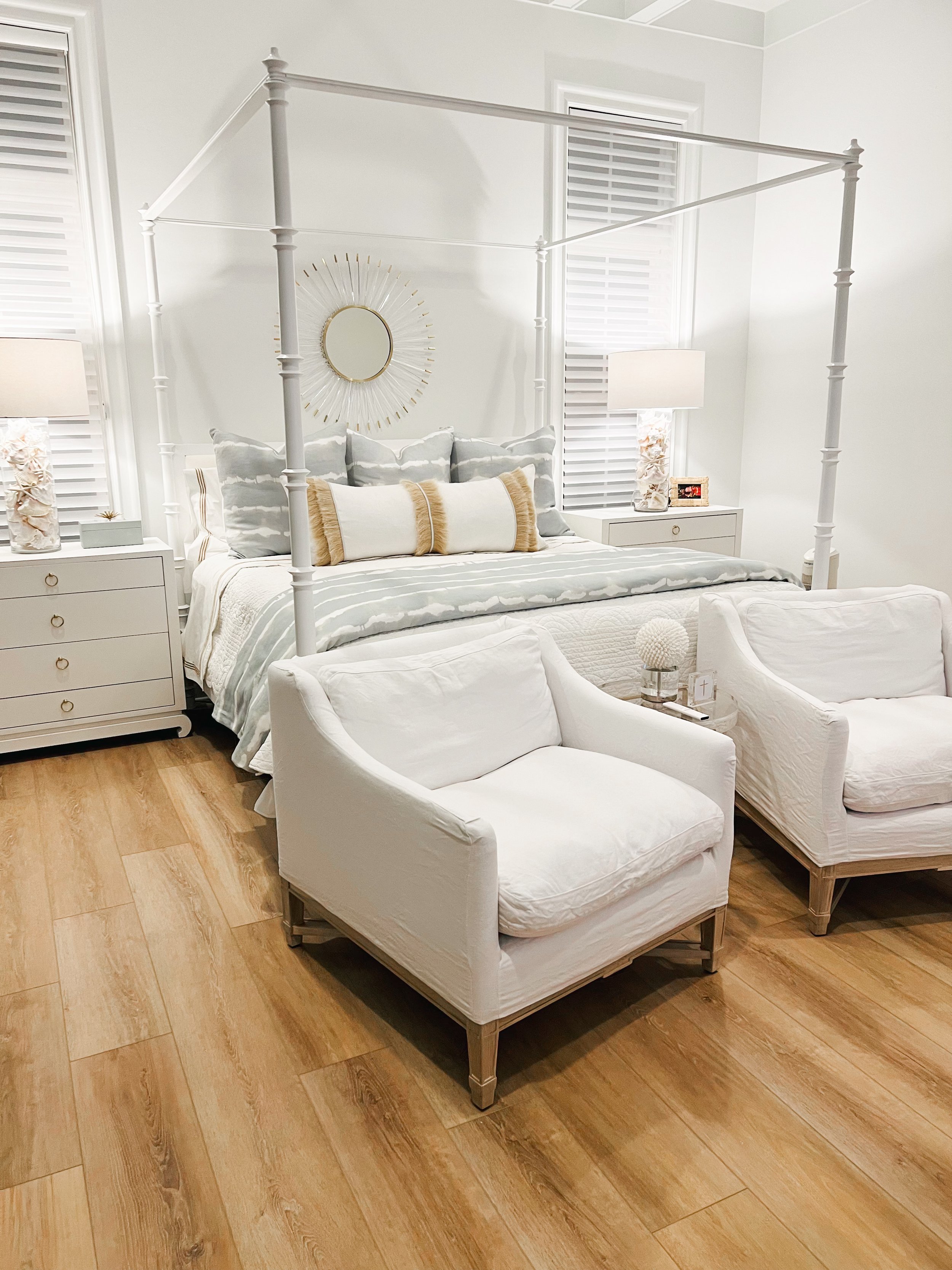 A stylish bedroom with a white canopy bed, light blue and white bedding, and two white armchairs on a wooden floor. The room features two bedside tables with lamps, and a decorative sunburst mirror above the bed.