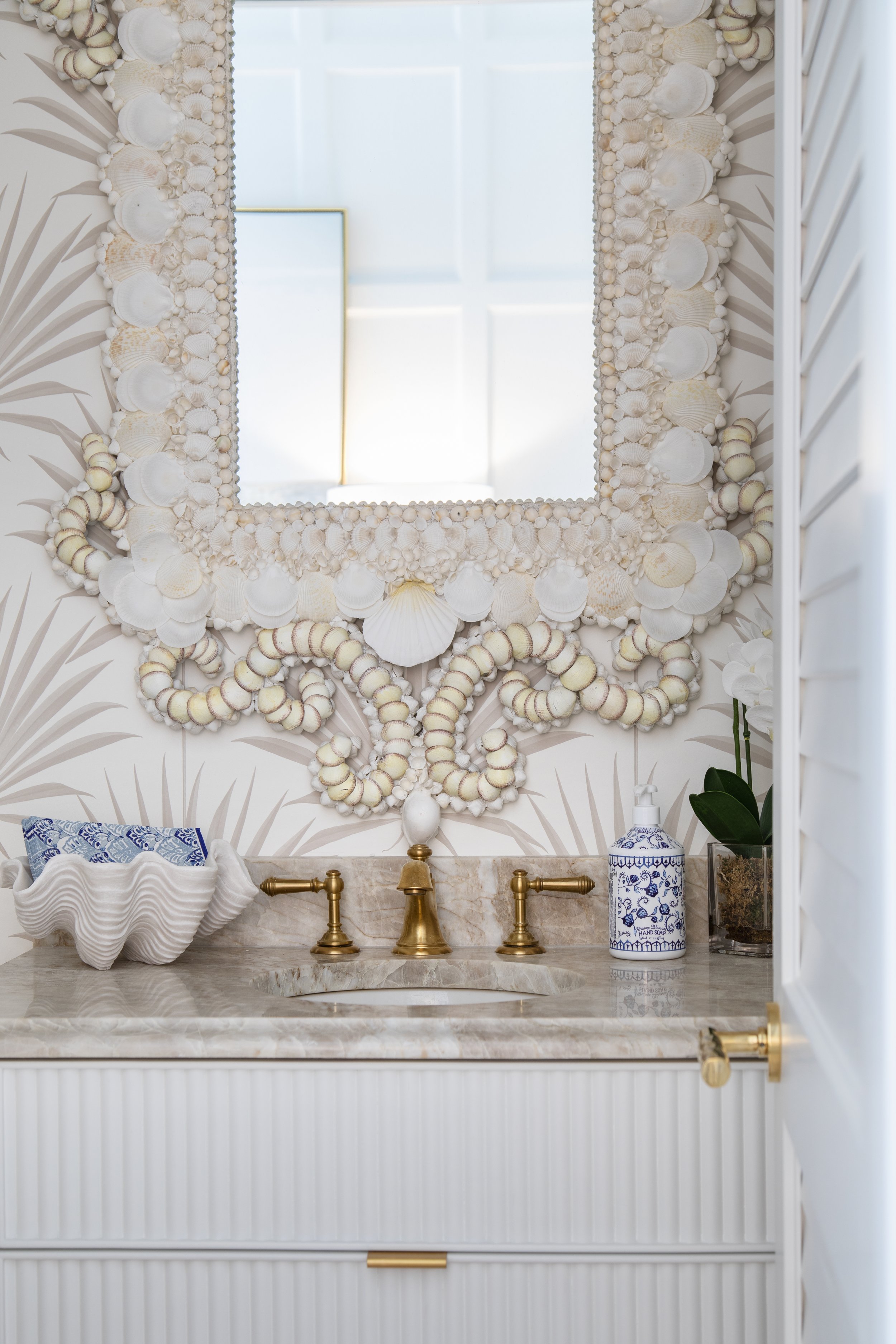 Bathroom vanity with shell-themed decor, intricate mirror, gold faucets, seashell-shaped dish, decorative soap dispenser, and orchid plant.