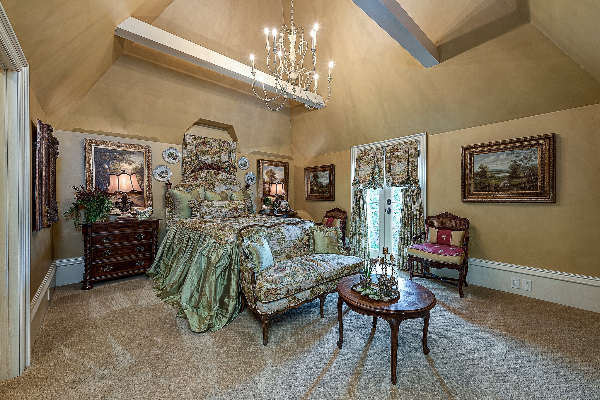 Elegant vintage bedroom with ornate chandelier, floral-patterned bedding, wooden furniture, wall paintings, and decorative pillows.