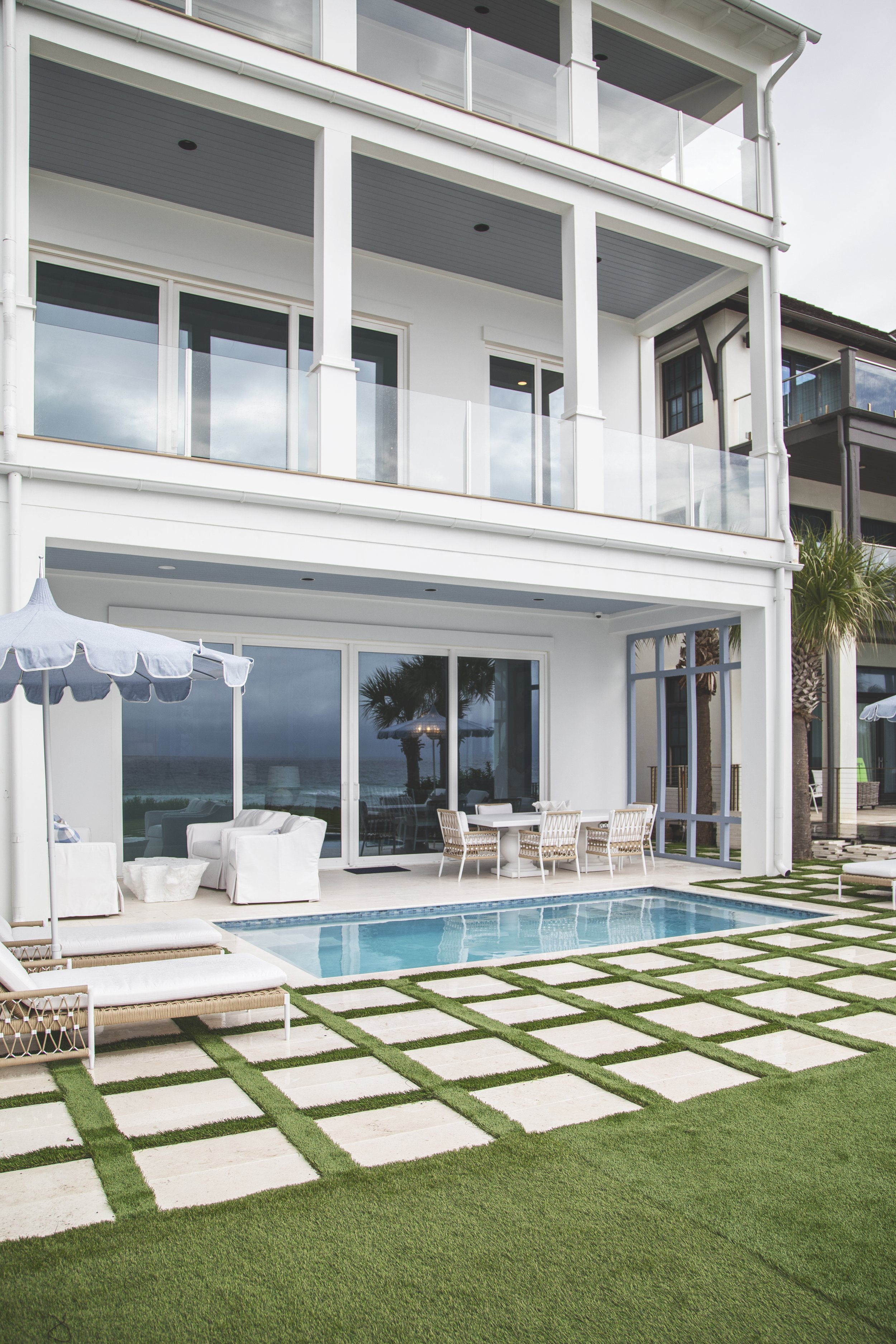 Modern white beachfront house with multiple balconies, glass railings, and a small pool surrounded by patio furniture and green lawn with stone tile pathway.