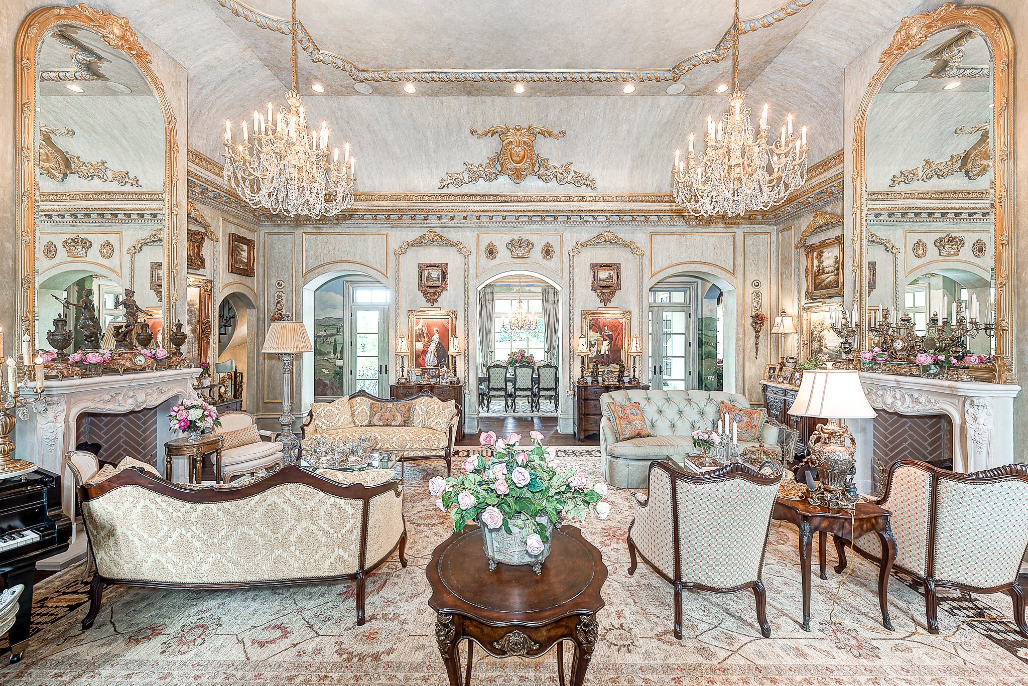 Luxurious living room with antique furniture, mirrored walls, crystal chandeliers, and ornate decor. Features a grand fireplace, upholstered chairs, floral arrangements, and intricate ceiling designs.