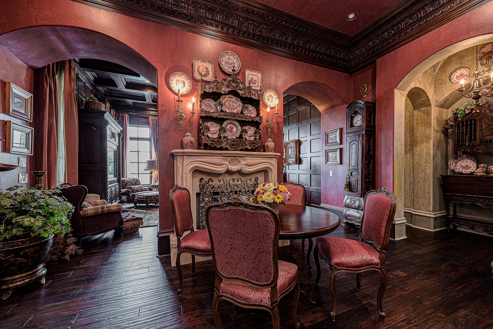 Luxurious traditional dining room with ornate red walls, antique furniture, and a decorative fireplace. Elegant chairs surround a round table, and decorative plates adorn the fireplace mantel. The room features rich wood flooring and ambient lighting