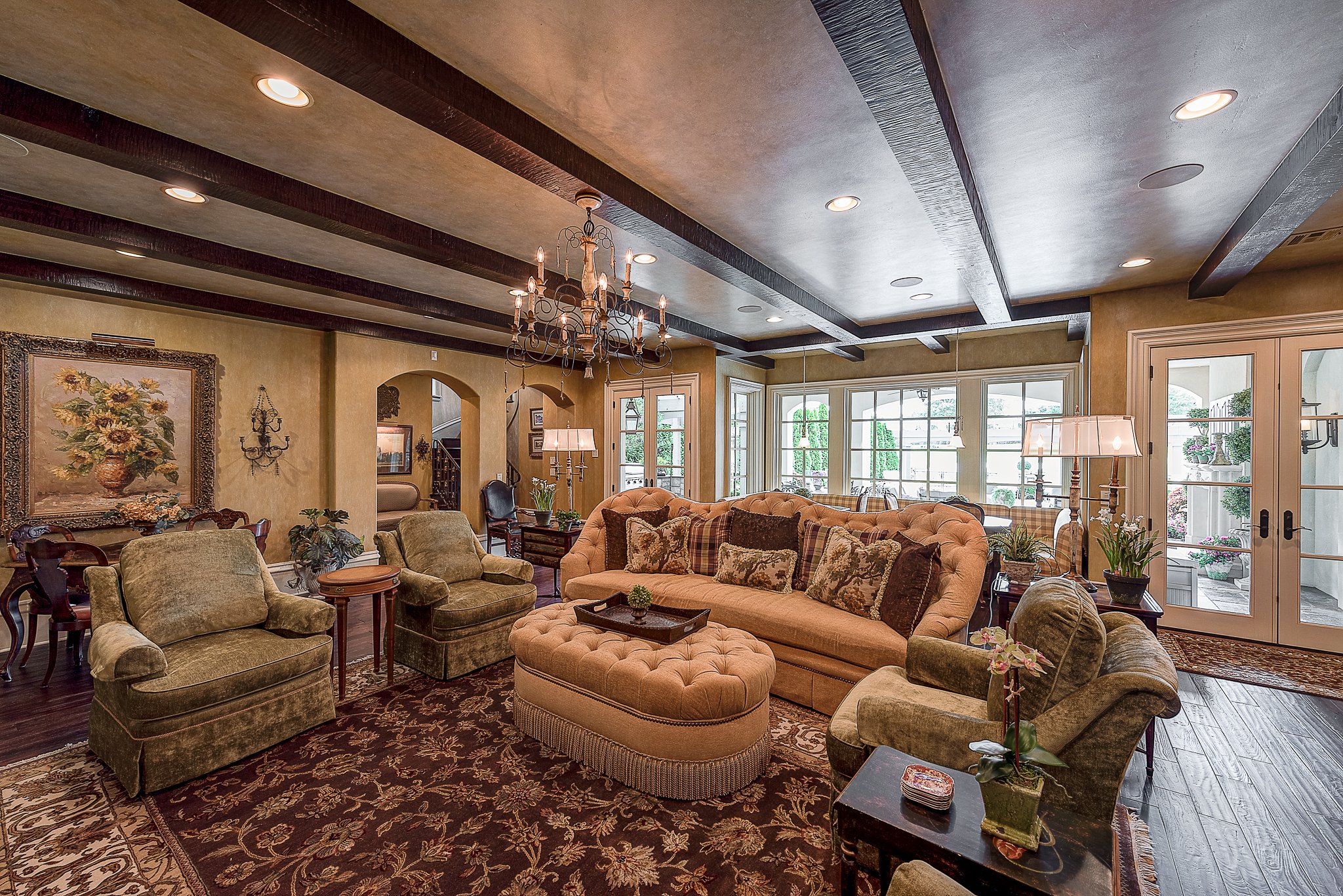 Luxury living room with plush seating, chandelier, and decorative rug, featuring large windows and a painting of sunflowers.