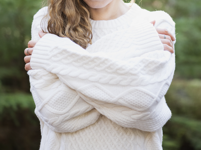 Person wearing a white cable-knit sweater with arms crossed, outdoors in a green environment.