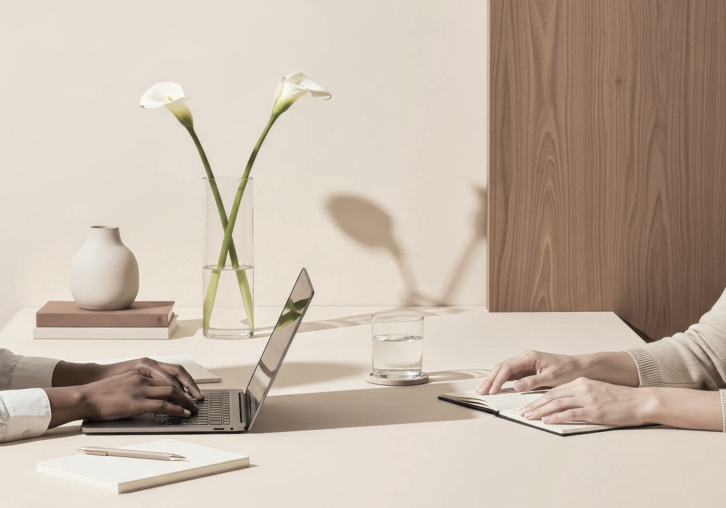 Two people sitting across a beige table, one typing on a laptop and the other holding an open notebook, with a glass of water, a vase with two white calla lilies, and a ceramic vase on stacked books in the background.