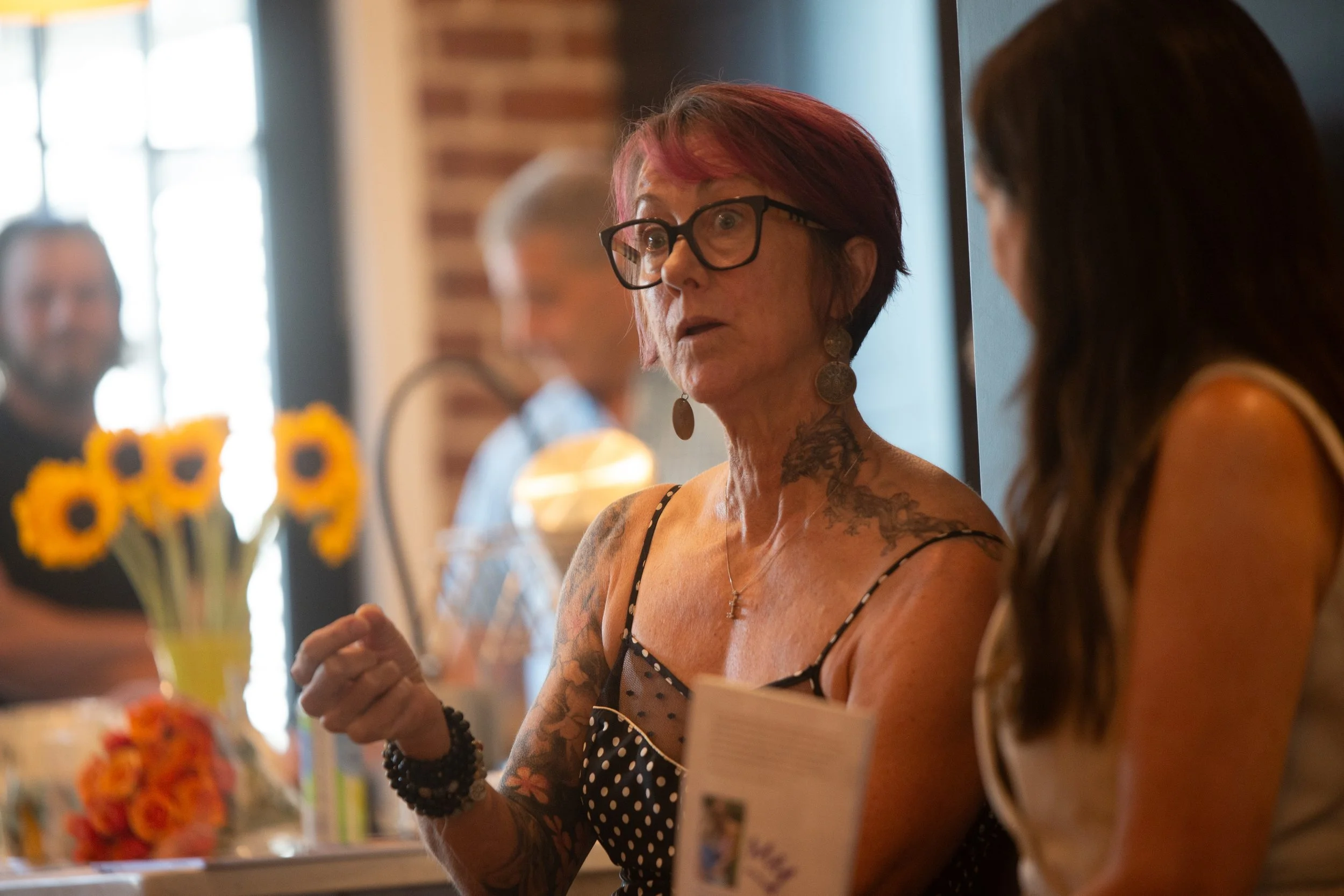 A woman with tattoos and glasses speaking to another woman in what appears to be a bakery or cafe, with people and flowers in the background.