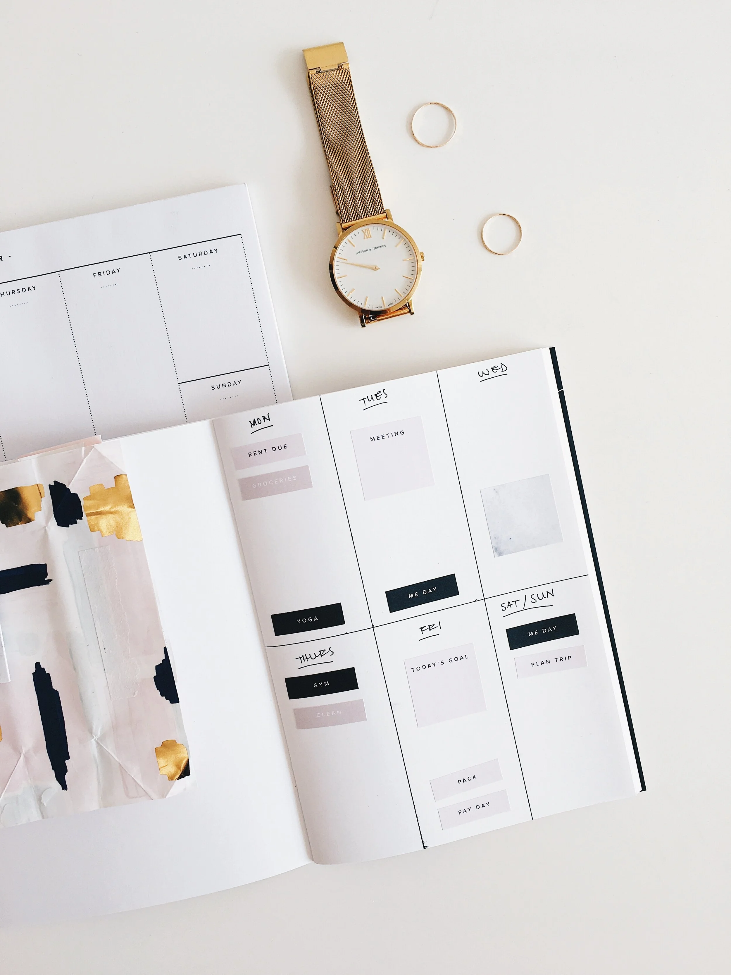 A weekly planner open on a white desk, with a gold watch, two rings, and some gold and black decorative paper nearby.