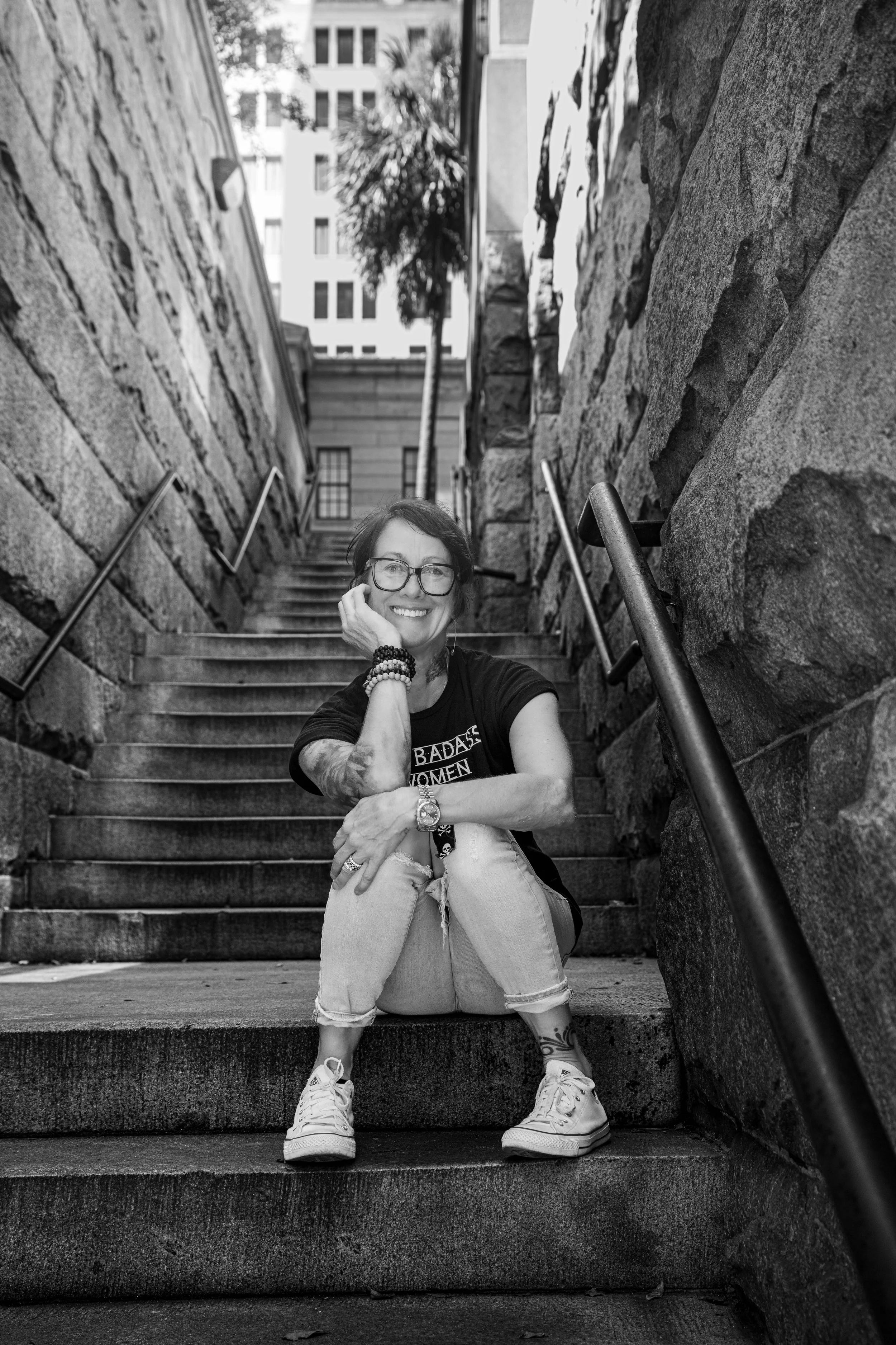 Smiling woman with glasses and tattoos sitting on outdoor stairs, wearing a black t-shirt and ripped jeans, surrounded by stone walls and metal railings, in an urban setting.