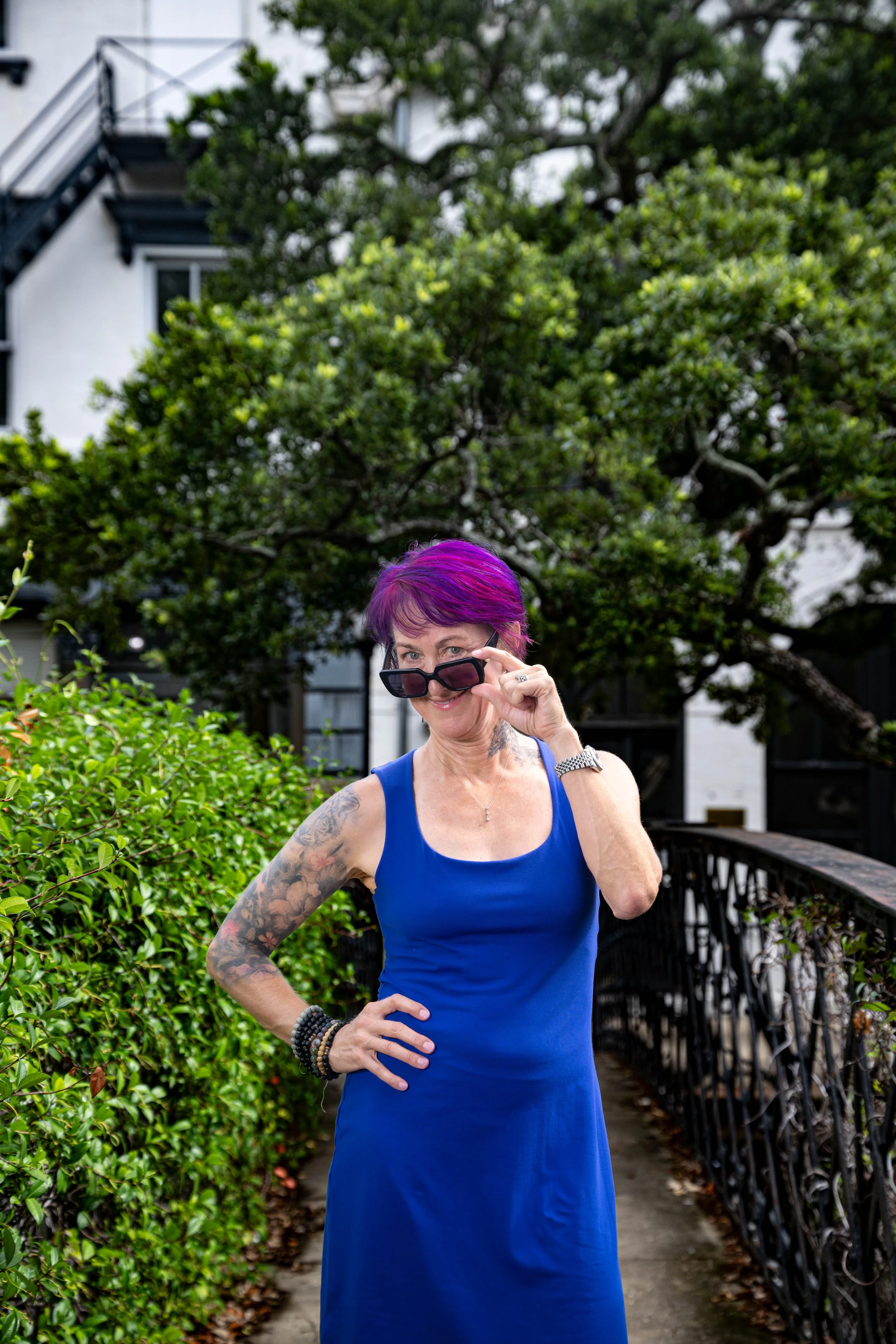 A woman with purple hair in a blue sleeveless dress lifting sunglasses and smiling on a walkway with greenery on both sides, in front of a house with trees in the background.