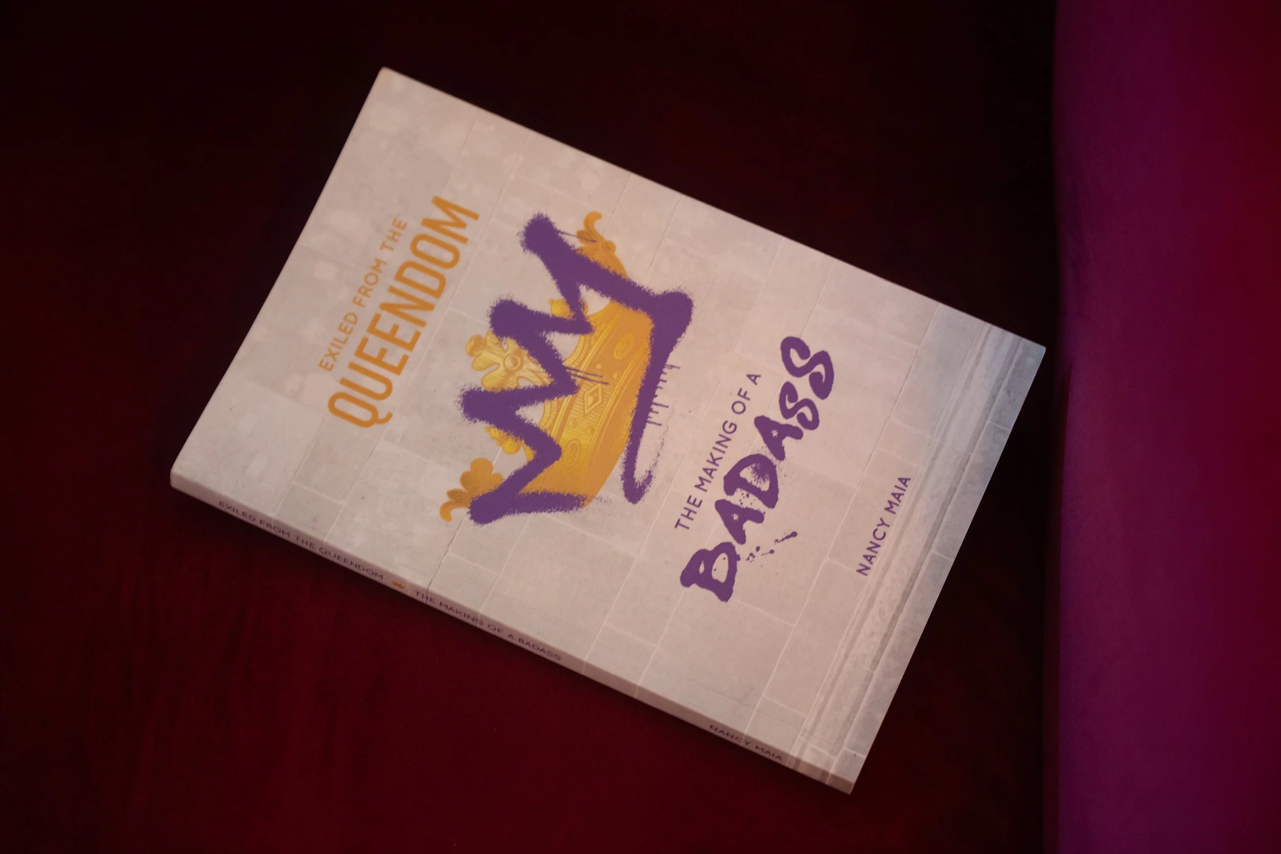 Book titled 'Exiled from the Queen's Court: The Making of a Badass' by Nancy Maia, with a purple and yellow crown illustration on the cover, placed on a dark surface.