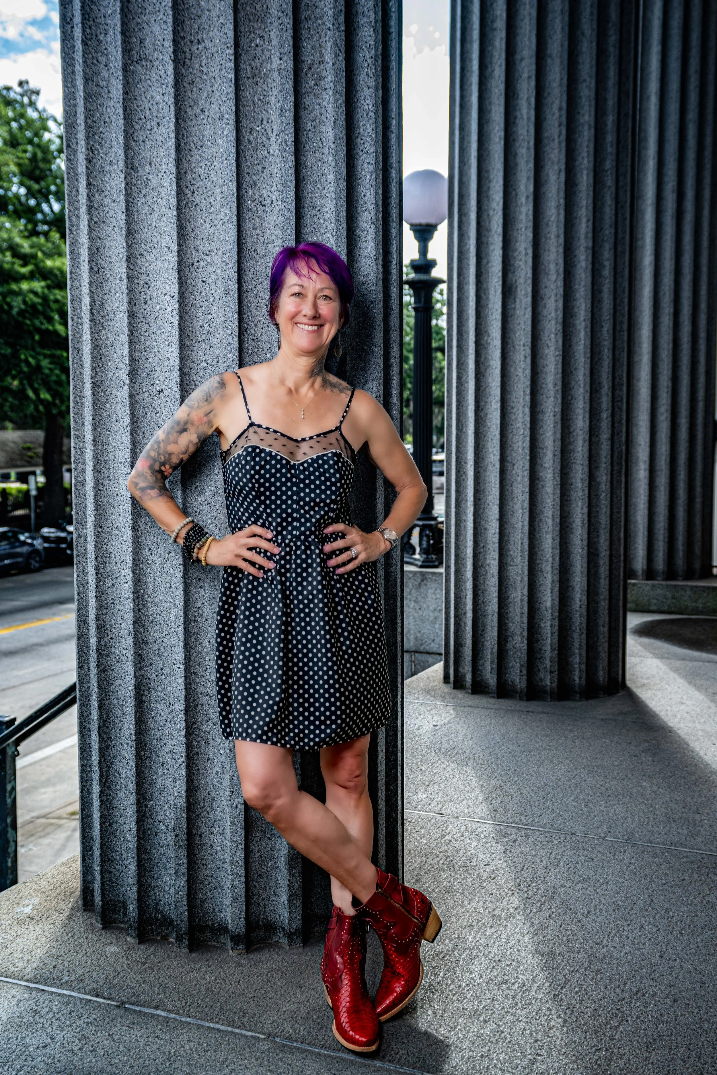 A woman with purple hair wearing a black polka dot dress and red boots standing outdoors near a building with columns.