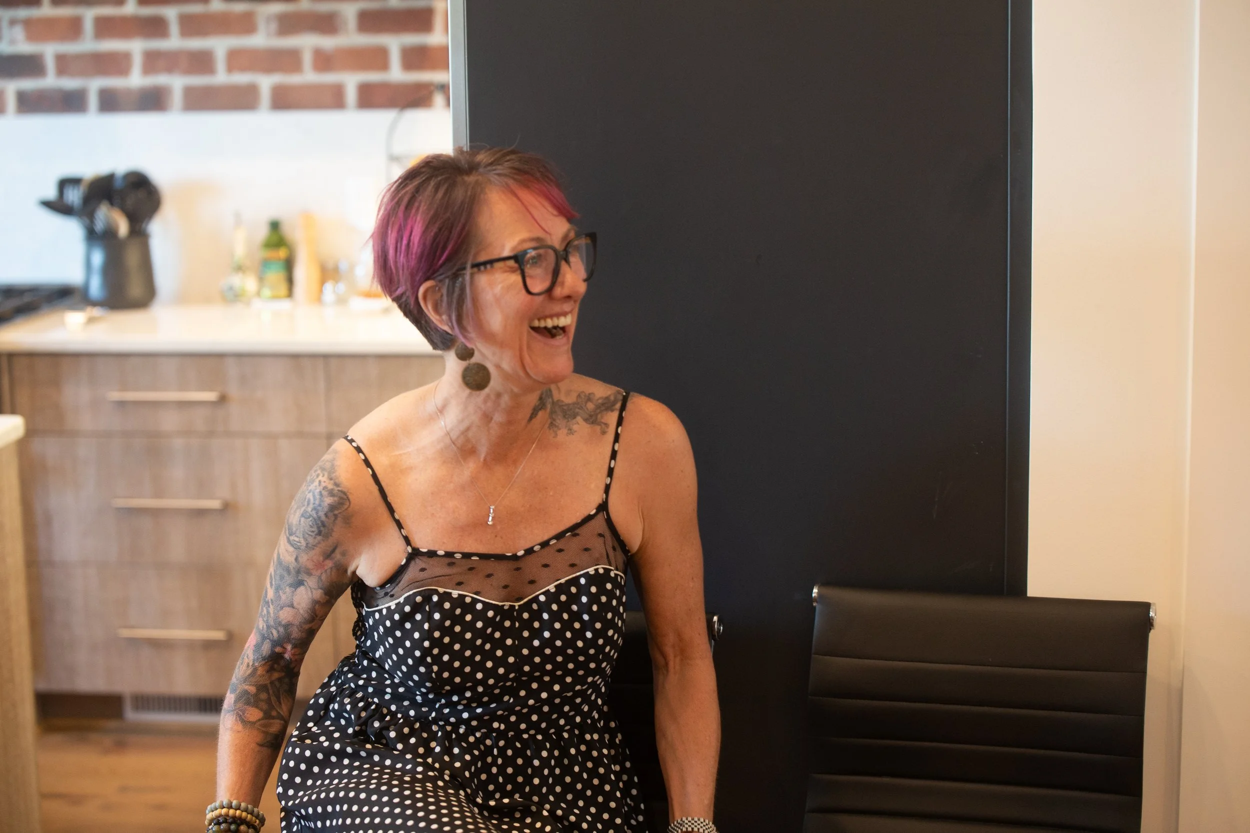 A woman with short, pink-tinted hair wearing glasses and a black polka dot dress, smiling and laughing in a kitchen setting.