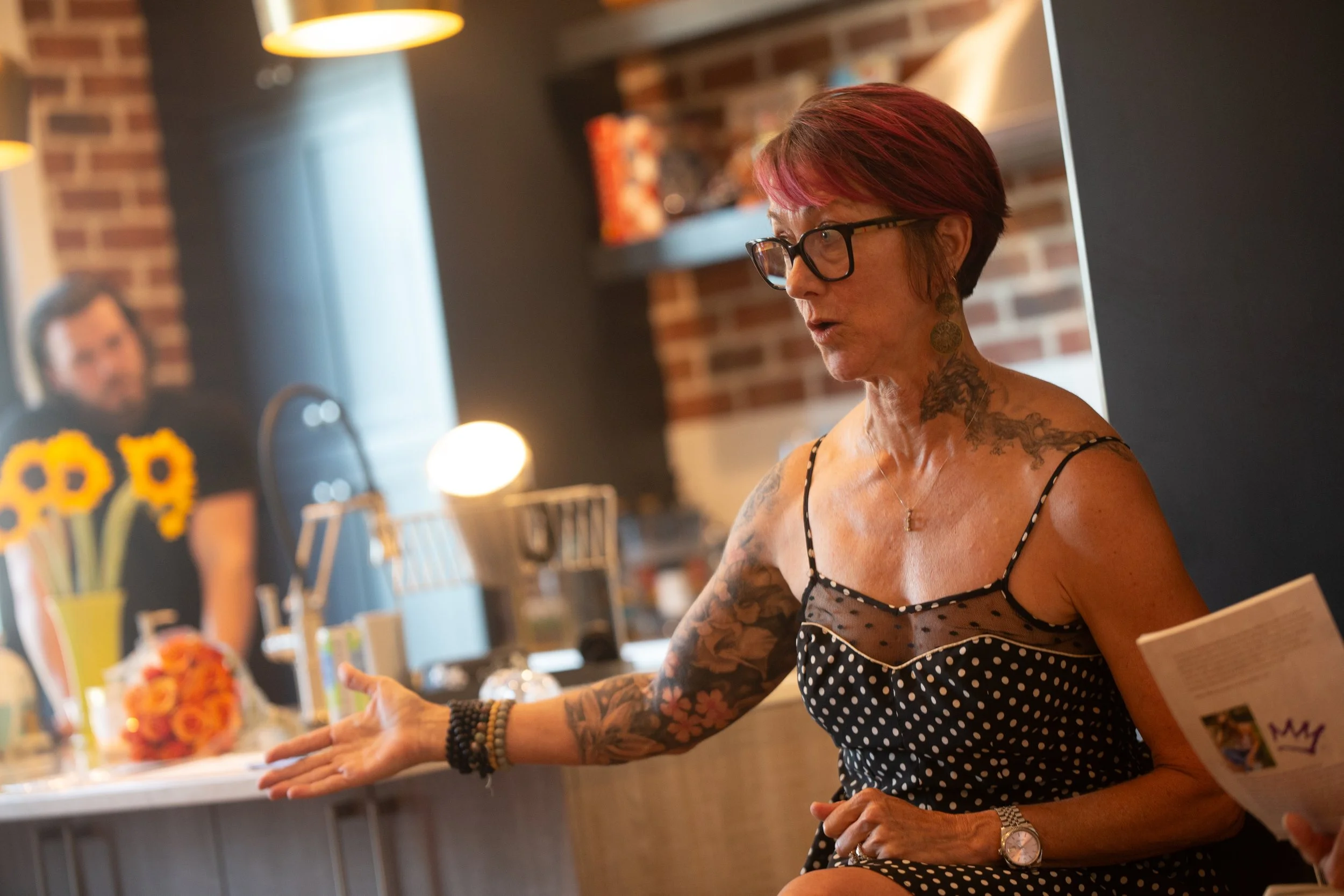 A woman with short red hair, glasses, and tattoos on her arms and neck is speaking and gesturing with her hand in a cozy kitchen environment. She is wearing a black polka dot dress with thin straps and is holding a piece of paper. In the background, a man with dark hair is blurred, standing near a counter with a bouquet of orange flowers and sunflowers on the kitchen island.