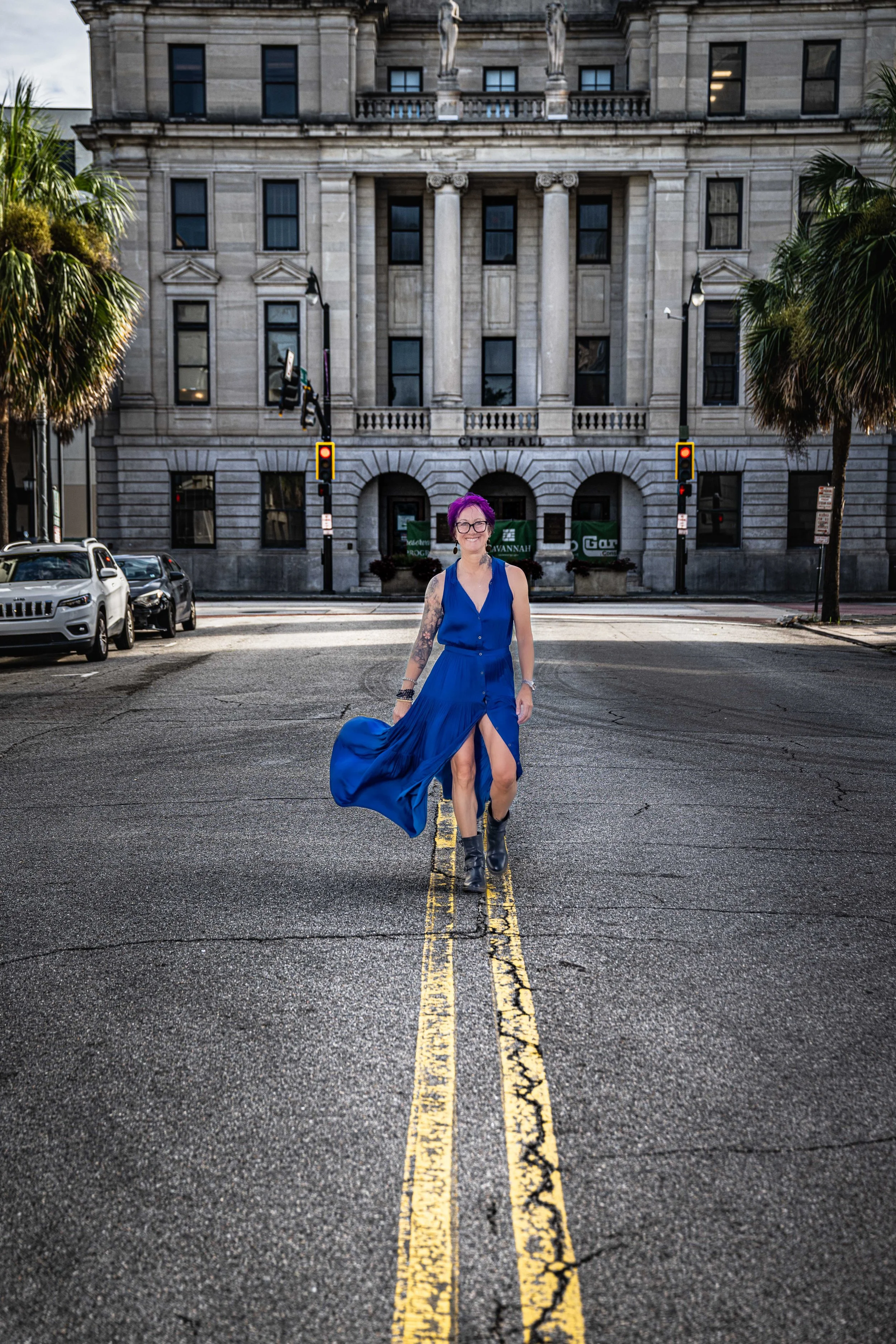 Woman with purple hair in a blue dress walking across the street in front of a city hall building, with cars parked on the side and palm trees on either side.