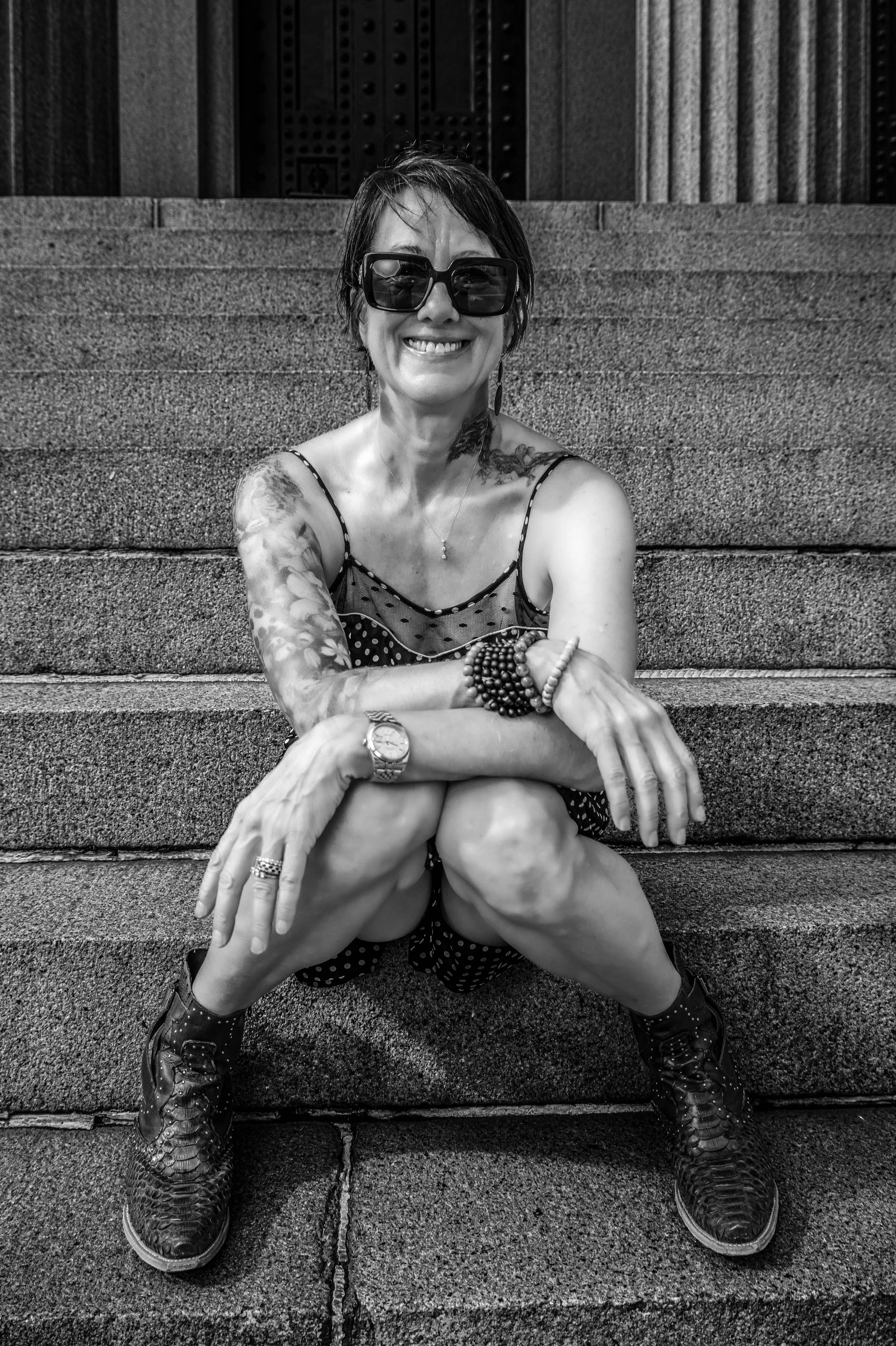 A smiling woman sitting on outdoor stairs, wearing dark glasses, a polka dot dress, and sneakers, with tattoos on her arm and multiple bracelets.