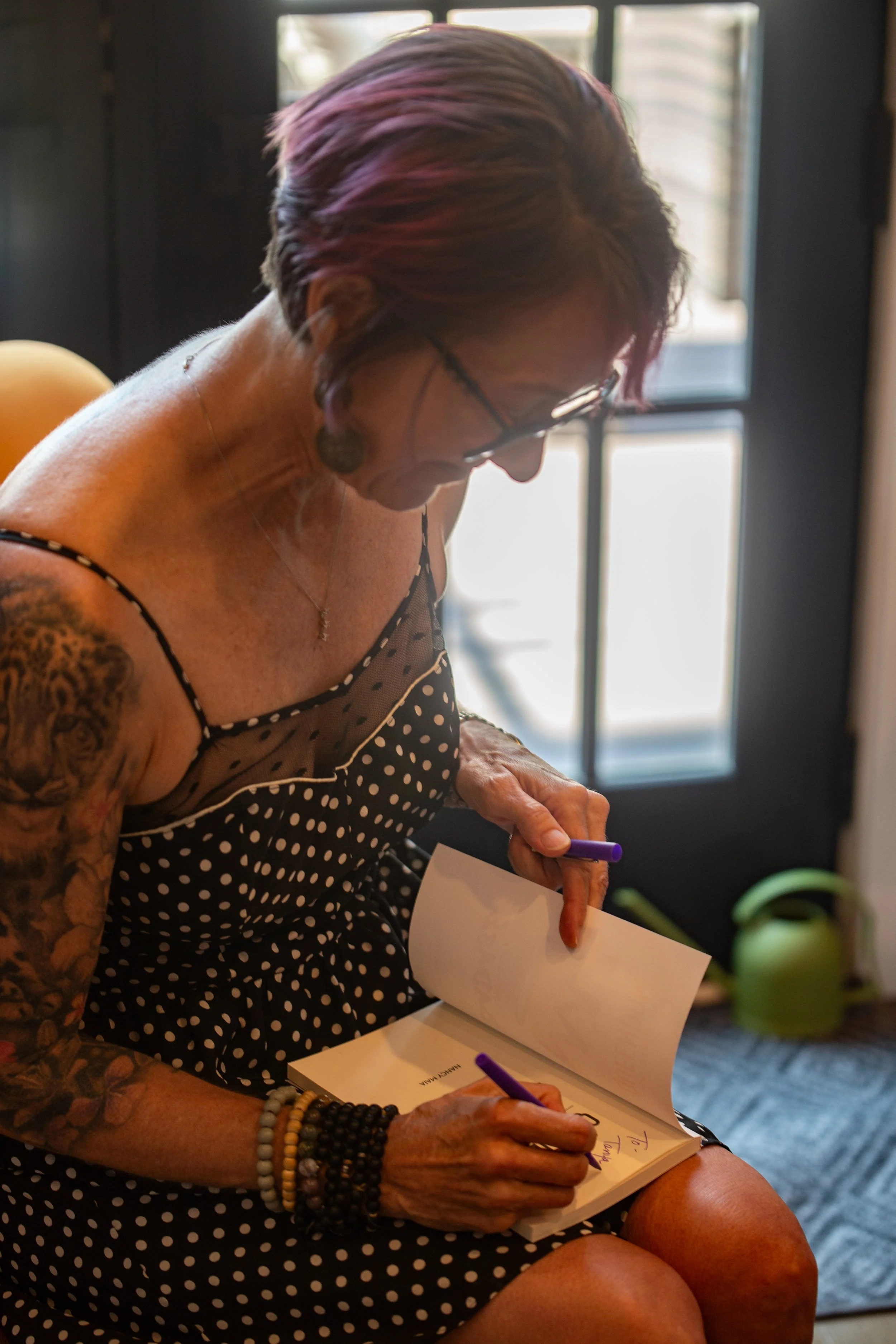 A woman with short, purple-tinted hair and glasses, wearing a black polka dot dress, sitting and writing in a book with a purple pen.
