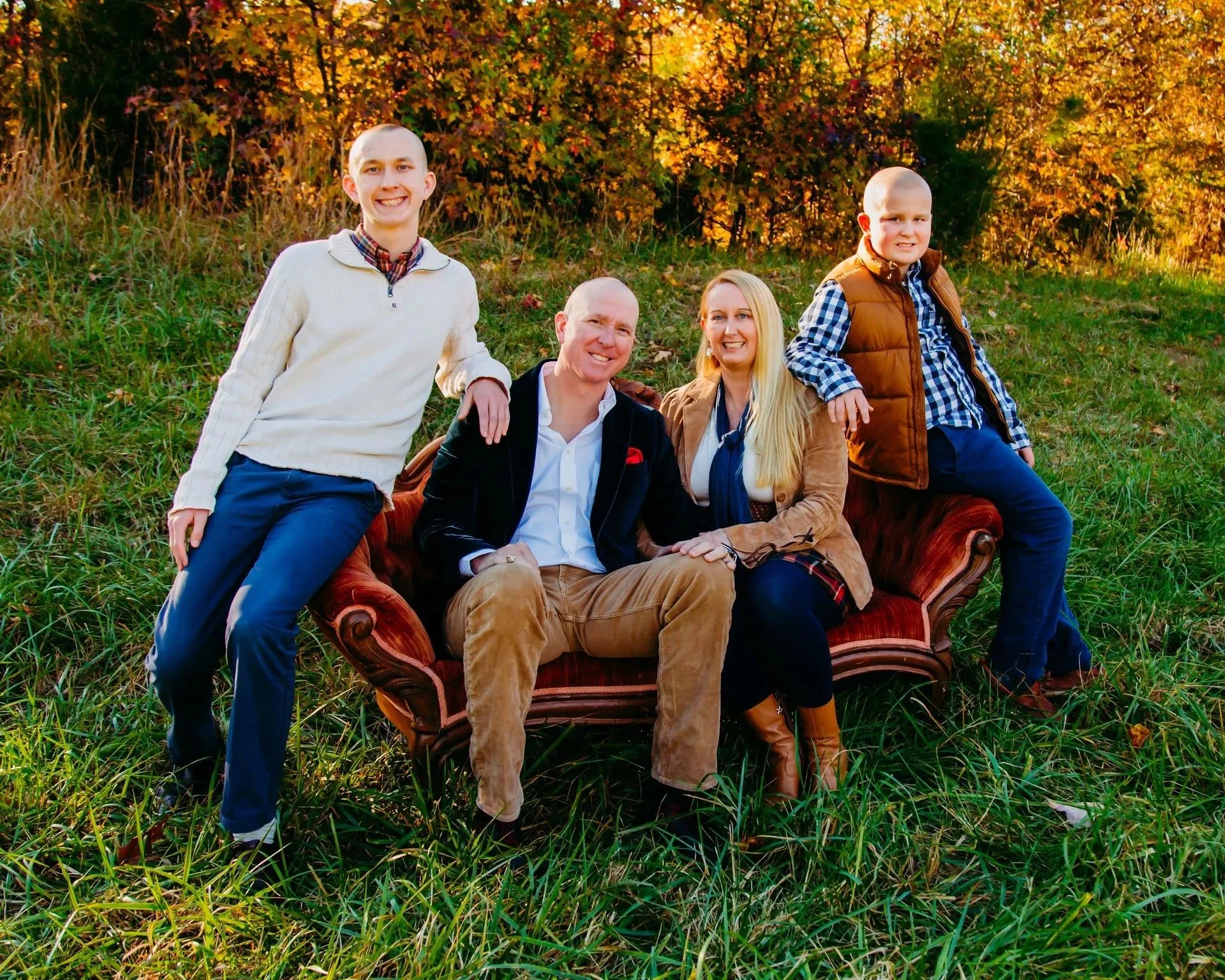 A family of five posing outdoors in a field with autumn trees in the background. They are sitting and standing around a vintage red velvet sofa, smiling in the sunlight.