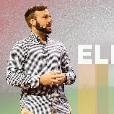 Man with a beard wearing a checkered shirt giving a presentation or speech, with a microphone headset, standing in front of a colorful gradient background.