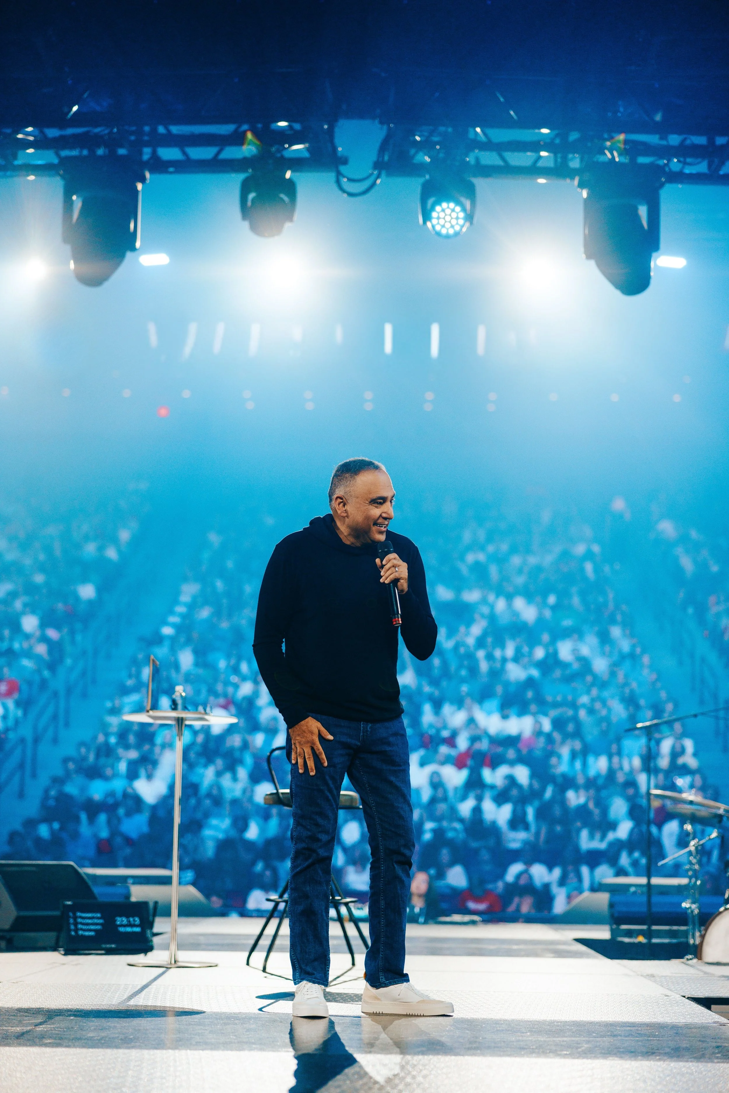 A man with short, slicked-back hair and a beard, wearing a dark button-up shirt, stands at a pulpit or podium with his hands raised mid-gesture, speaking or presenting in a dimly lit room with a large screen in the background.
