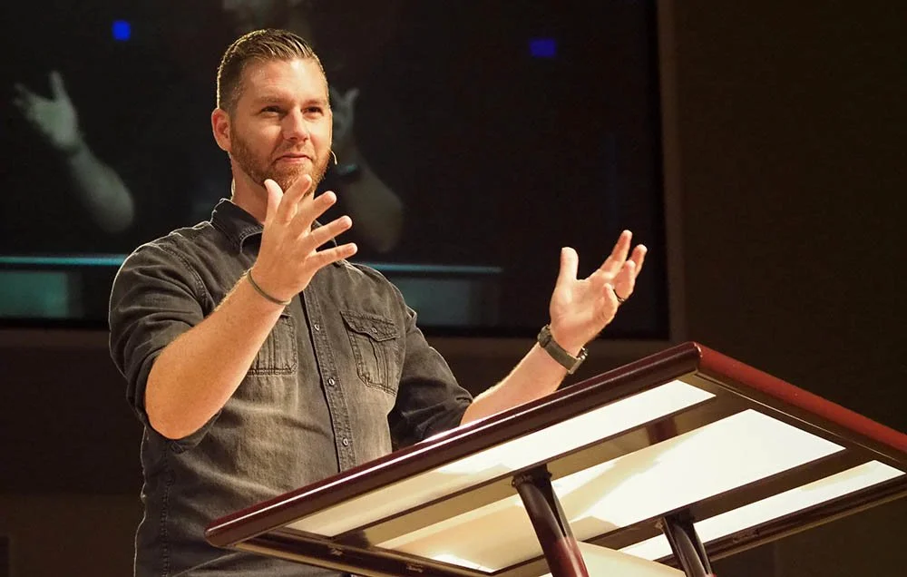A man with short, slicked-back hair and a beard, wearing a dark button-up shirt, stands at a pulpit or podium with his hands raised mid-gesture, speaking or presenting in a dimly lit room with a large screen in the background.