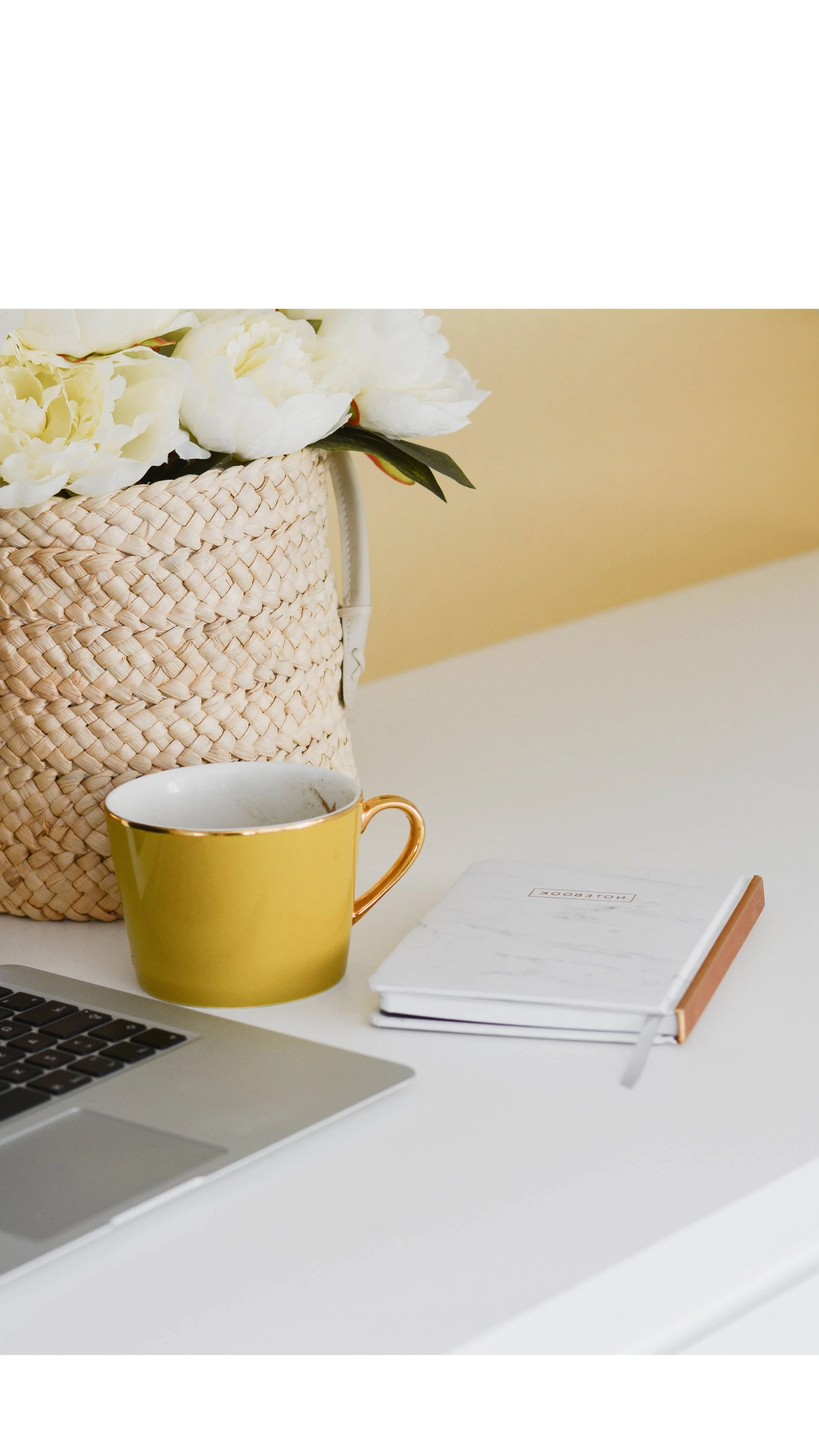 A white desk with a yellow mug, a laptop, a closed notebook, and a woven basket with white flowers sitting on top, against a yellow wall background.