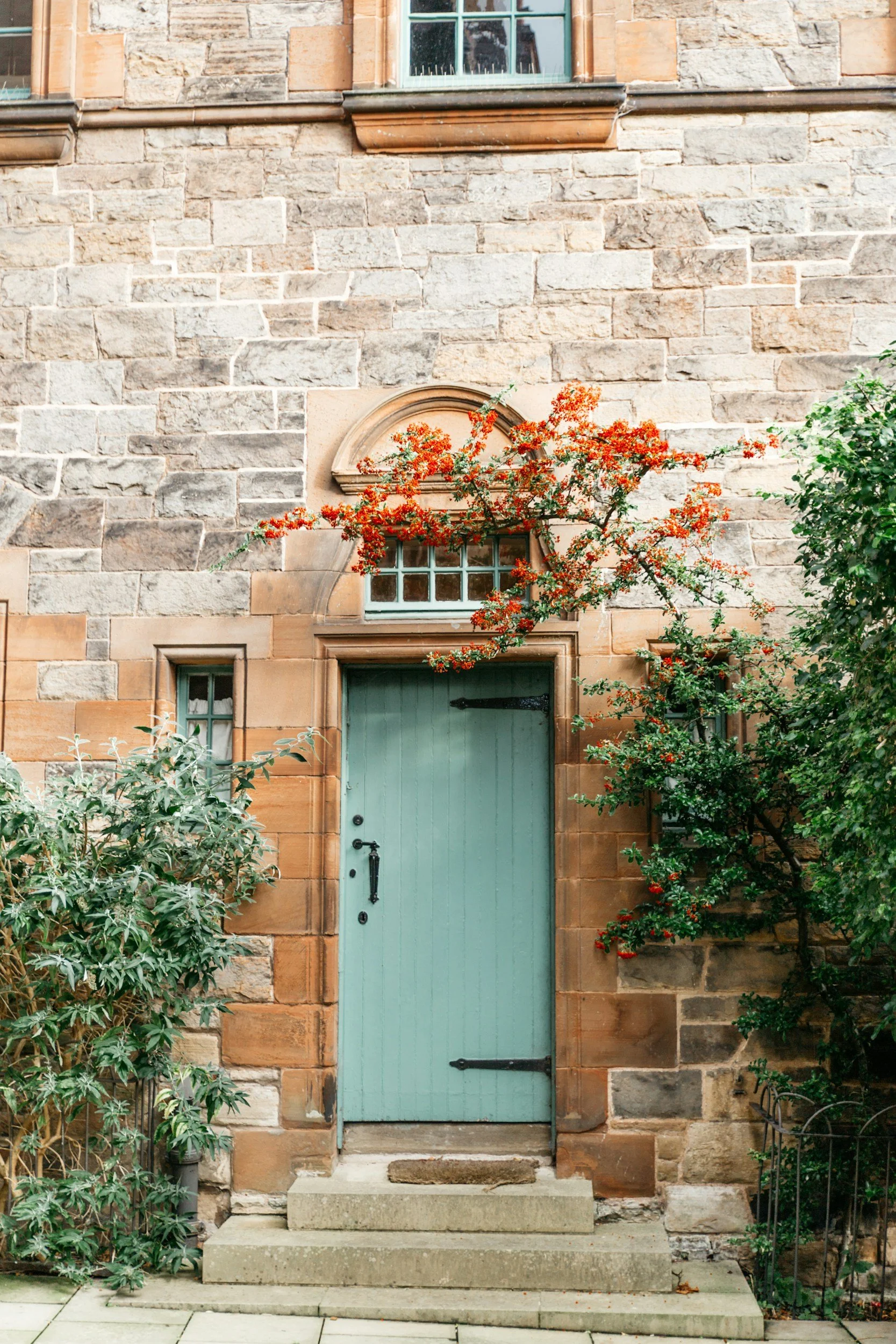 A historic stone building with a light blue wooden door, small rectangular windows above the door, and a climbing plant with red berries growing near the entrance.