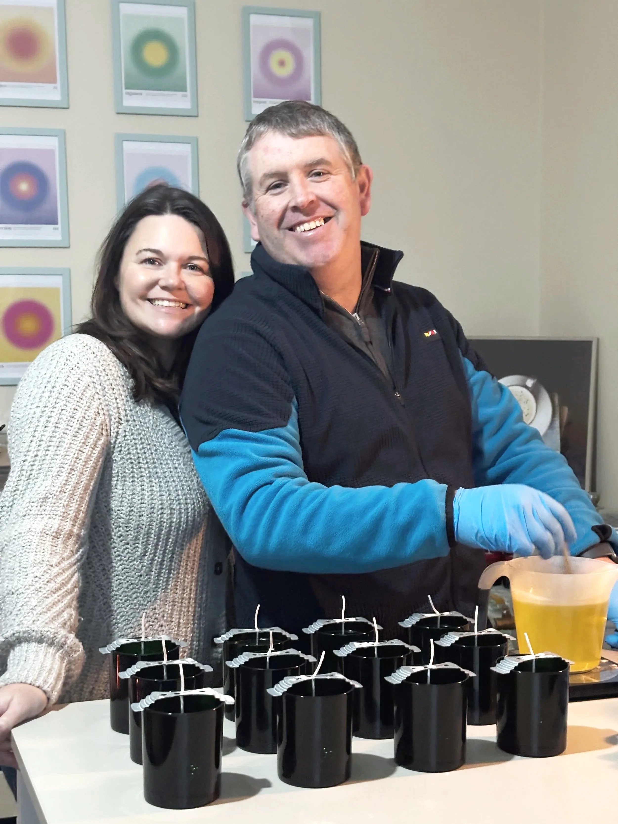Two people smiling at a table with black candles and a pitcher of yellow liquid, with colorful abstract art on the wall behind them.
