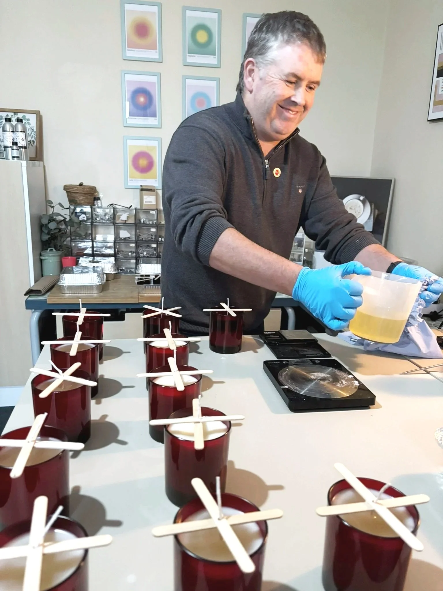 A man wearing blue gloves is pouring a yellow substance into clear cups on a table, with several red cups with white sticks standing on the surface. The scene appears to be related to a scientific or culinary activity.
